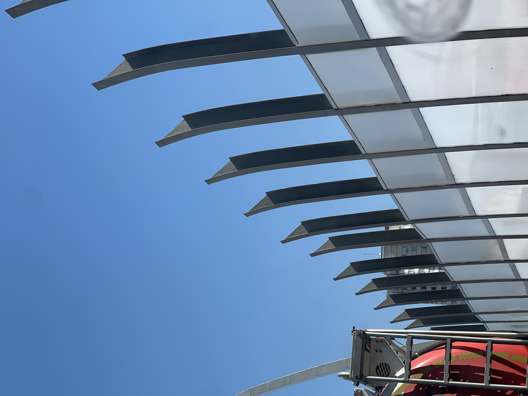 Here's a concise description of the image:
The image presents an upward view of a modern architectural structure against a clear blue sky. The structure consists of a series of parallel, horizontal, gray metal beams with angled edges. These beams are spaced with transparent or translucent panels between them, creating a canopy-like effect. The structure appears to be part of a building or perhaps a pedestrian walkway. In the lower-right corner of the image, a portion of a red object with a metallic frame is visible, possibly part of a mechanical device or vehicle. In the background, there is the outline of a building, perhaps a business office. Based on the architecture, location and style, this image depicts Zaryadye Park, near the Red Square, in Moscow, Russia.