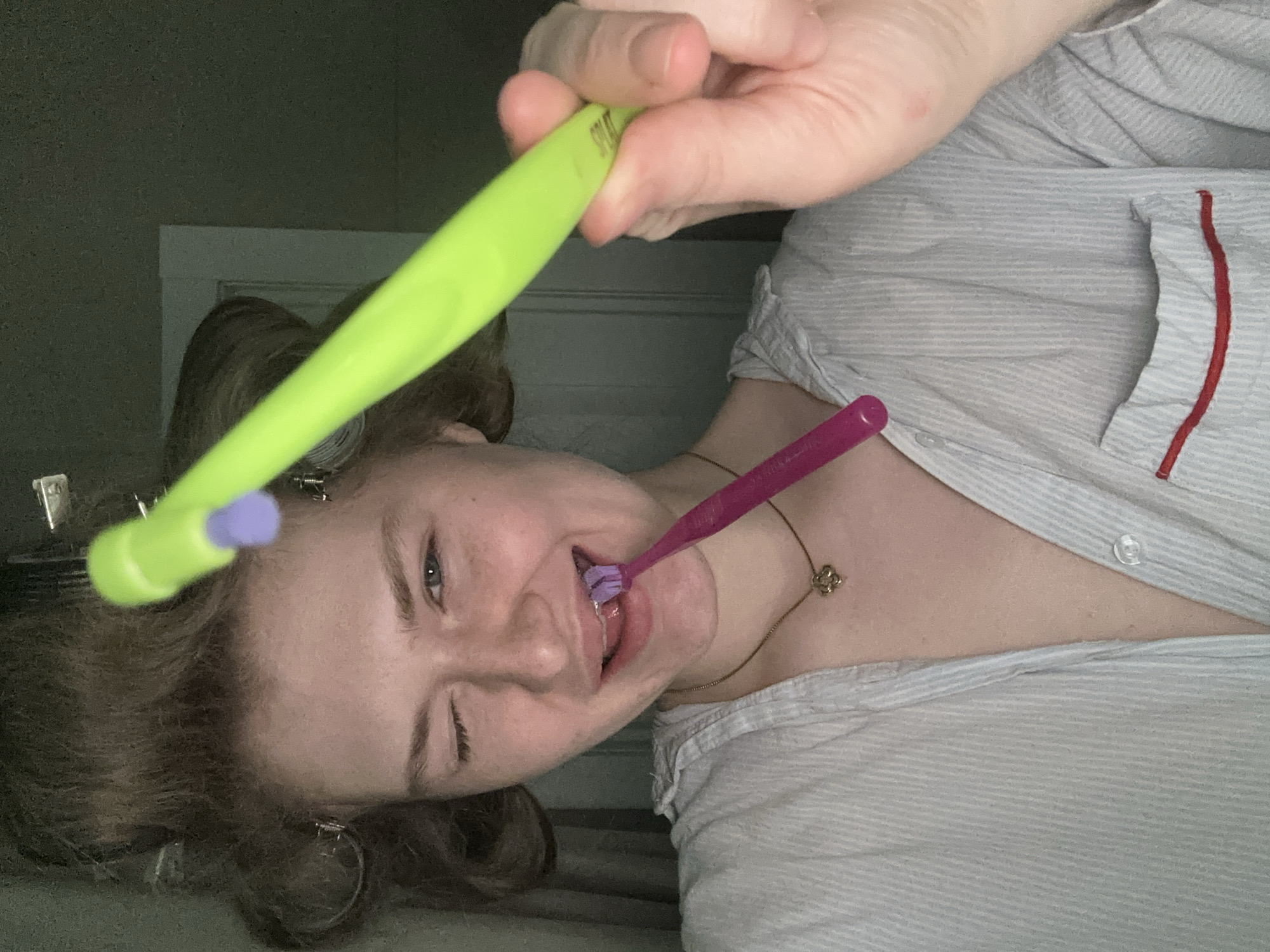 A woman is brushing her teeth with two toothbrushes, one purple and one neon green. She is wearing a striped shirt with red trim. Her hair is in curlers. She is also wearing a necklace. The location of the image is not discernible.