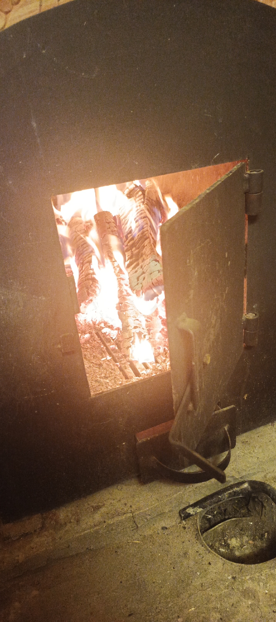 The image captures a close-up view of a burning fire within a large, dark-colored structure, likely a stove or furnace, located in Unknown, Unknown. A rectangular opening in the center reveals vigorously burning logs, emitting bright orange and red flames and glowing embers. To the right of this opening, a dark metal door, hinged on the right with two visible cylindrical hinges, is partially open, angled away from the main structure. The surrounding surface of the stove is a dark, matte material, possibly metal or stone, showing some texture and minor surface imperfections. The immediate foreground consists of a gritty, light-colored concrete floor, subtly illuminated by the firelight. In the lower right of the frame, a dark, curved metal object, possibly a tool handle or part of a grate, rests on the floor. Next to it, a circular opening in the concrete contains dark ash or debris and a dark, curved metallic edge.