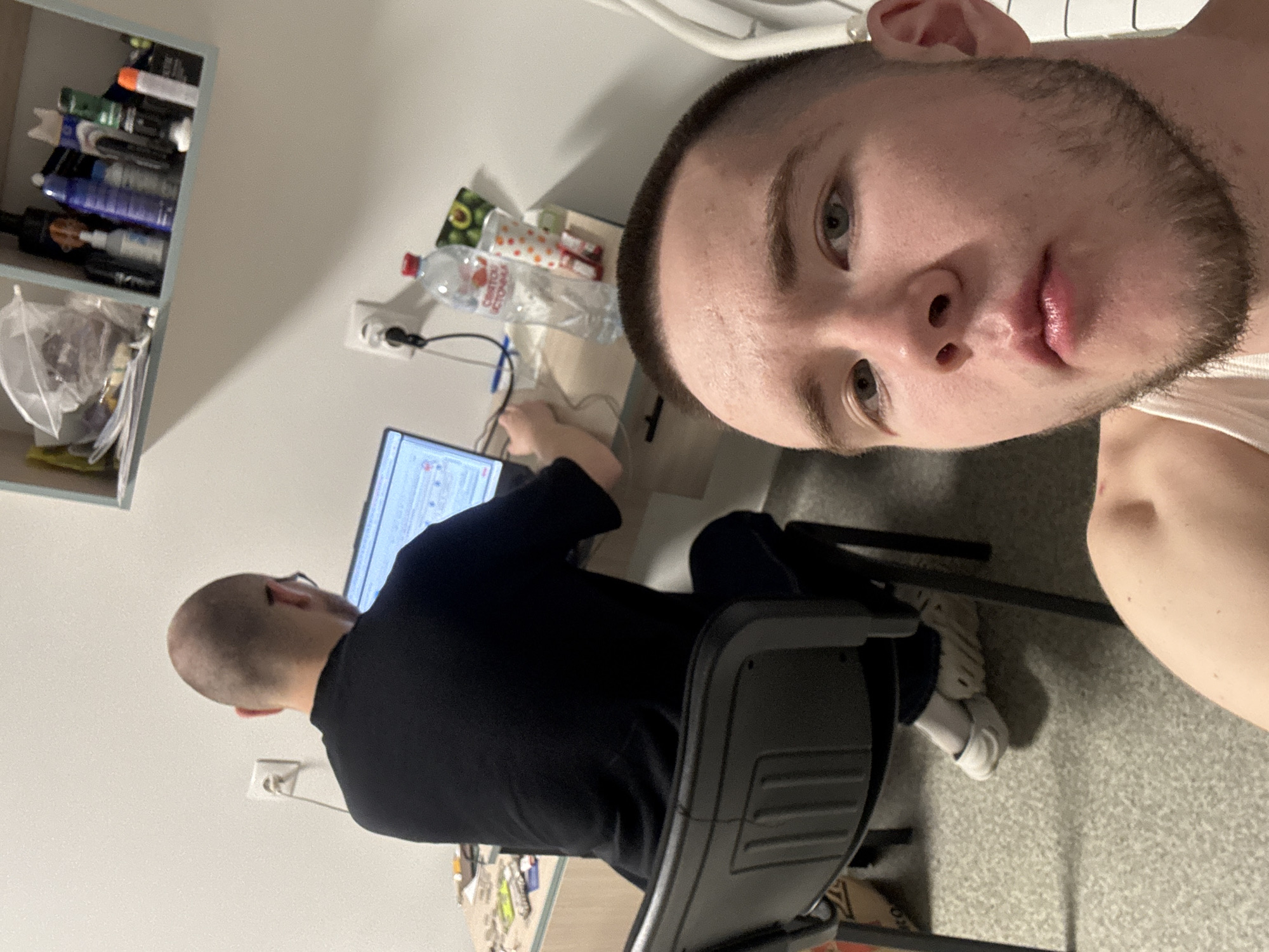The image depicts an indoor scene featuring two men. In the foreground, a man takes a selfie, looking directly into the camera with a neutral expression. He has a buzz cut, light-colored eyes, and light stubble on his face, wearing a white tank top.
In the background, a second man is seated at a desk, focused on a laptop screen. He is seen from behind, appearing bald or with very short hair, and wears a dark long-sleeved shirt and glasses. The laptop screen displays what appears to be a graphical user interface with text and elements.
The setting includes a light-colored wall with a power outlet, a grey carpeted floor, and a grey/blue shelving unit to the left. The shelves hold various items, including personal care products and a plastic bag. On the desk, next to the working man, there is a clear plastic water bottle with a red cap, labeled in Russian as "Святой Источник" (Svyatoy Istochnik), a pen, and a small package. The room appears functional, suggesting a workspace or living area, consistent with the location context of Kazan, Russia.