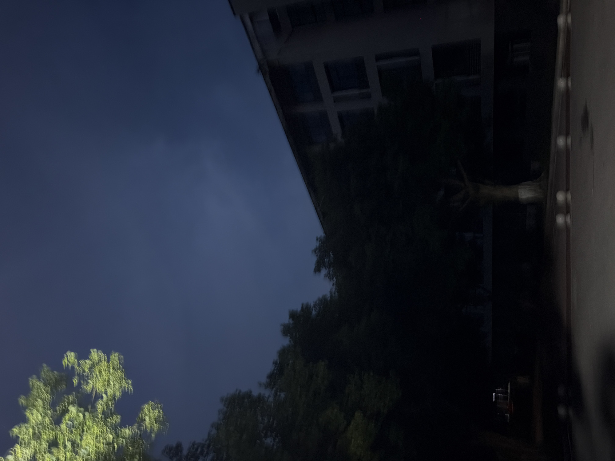 The image depicts a dark outdoor scene at night, possibly in the city of Jiangjin, China. A building with multiple windows is visible on the right side of the frame, partially obscured by trees. The sky is dark blue with some faint cloud cover. In the foreground, there is a paved area with what appear to be decorative bollards. The overall impression is of a quiet, possibly residential or institutional area at night.