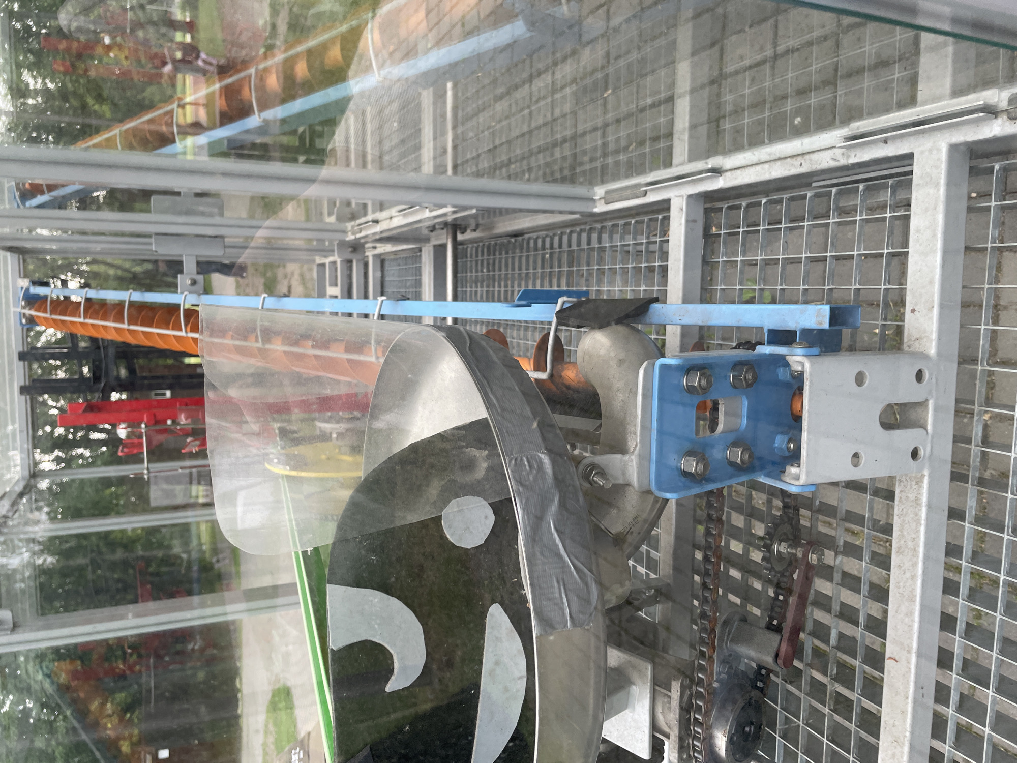 A still indoor shot shows a Rube Goldberg machine housed in a glass enclosure. The contraption features a series of blue and gray metal supports, a chain drive, and a spinning disk with a cartoon face. The enclosure sits on a metal grid platform. Through the glass, a park-like setting with greenery and other structures is visible. The city is not identifiable.