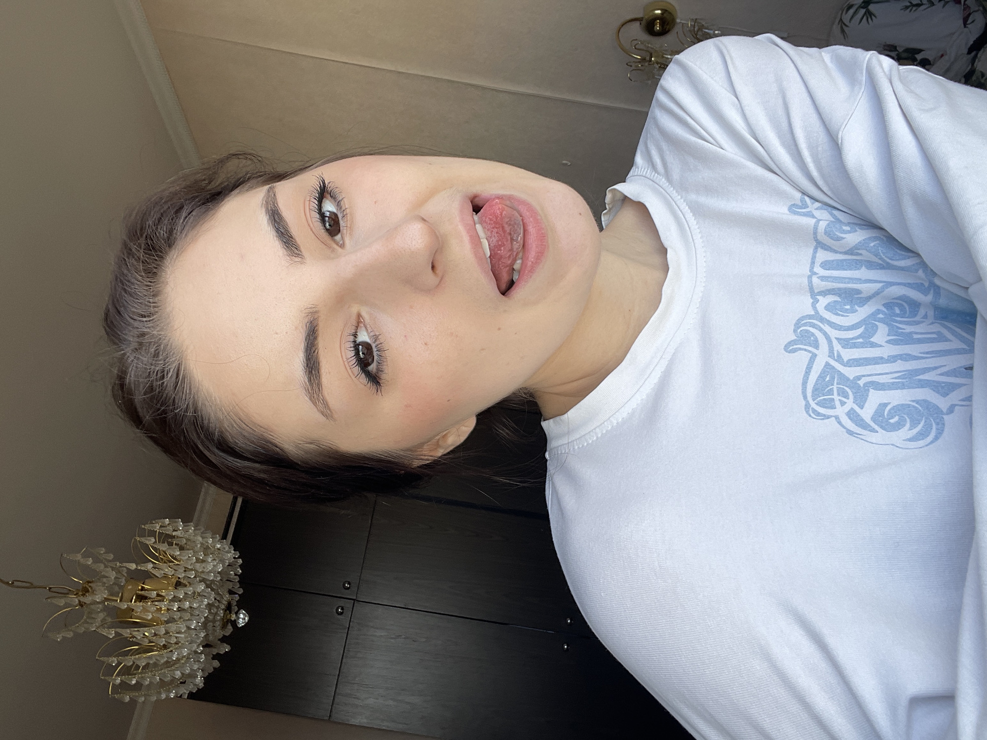 A young woman is posing for a photo in what appears to be an indoor setting. She is wearing a white long-sleeved shirt with a blue graphic. Her mouth is open, and her tongue is slightly extended. In the background, there is a dark cabinet or wall with metallic knobs. Above the woman and to the side is a chandelier with ornate gold-colored metal and clear decorative beads. The setting cannot be verified as Moscow, Russia, based on the provided image.