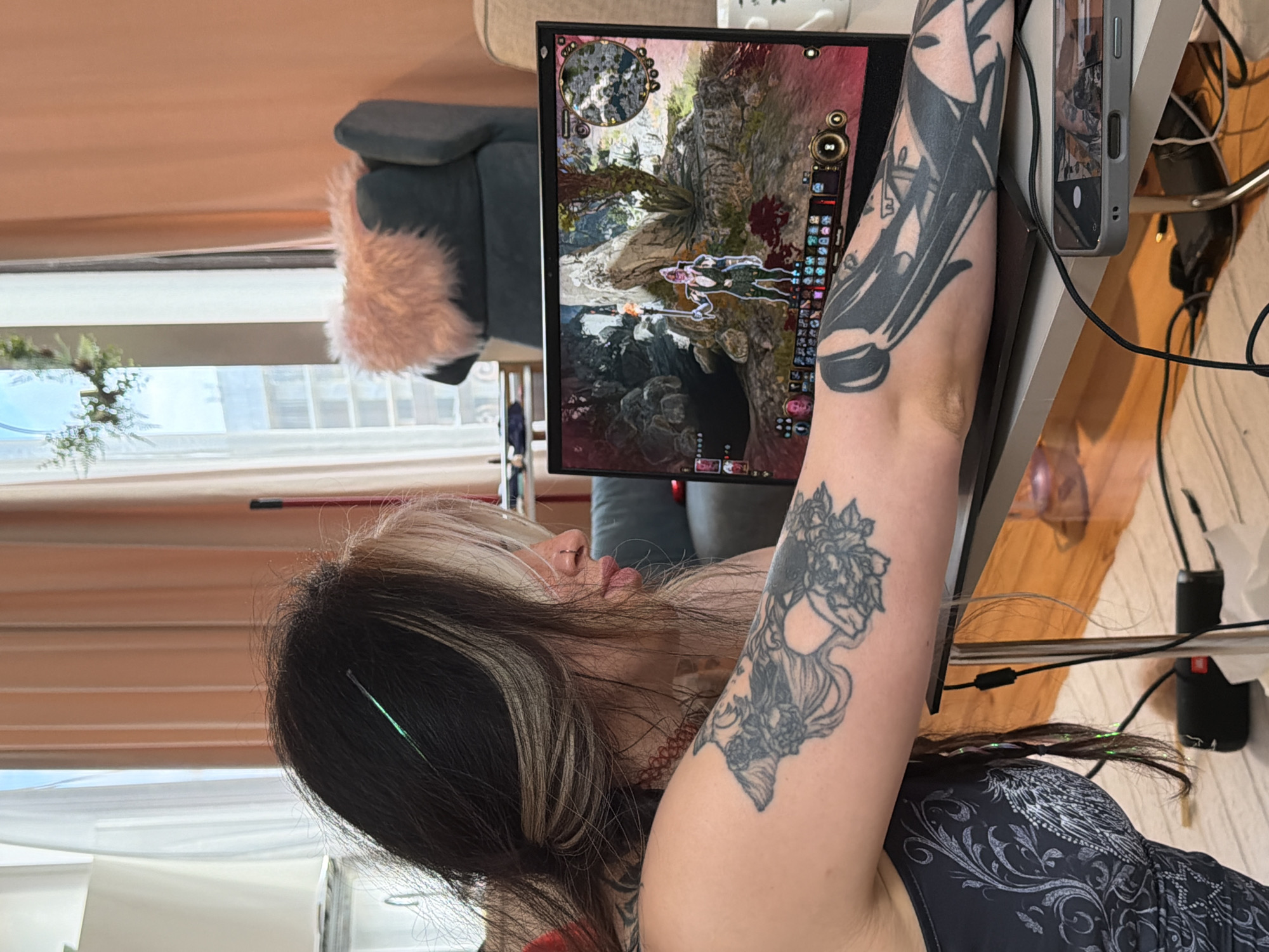 A person is seated in front of a computer monitor displaying a video game. The game appears to be set in a fantasy landscape with mountainous terrain, a character, and game interface elements visible on the screen. The person has dark hair with blonde streaks, and is wearing a black tank top. The person has two tattoos on their left arm. The person appears to be holding a cell phone in their left hand. The background includes a window, a chair with a pink pillow, and computer accessories on a desk.
There is no evidence to suggest a location in Budva, Montenegro.
