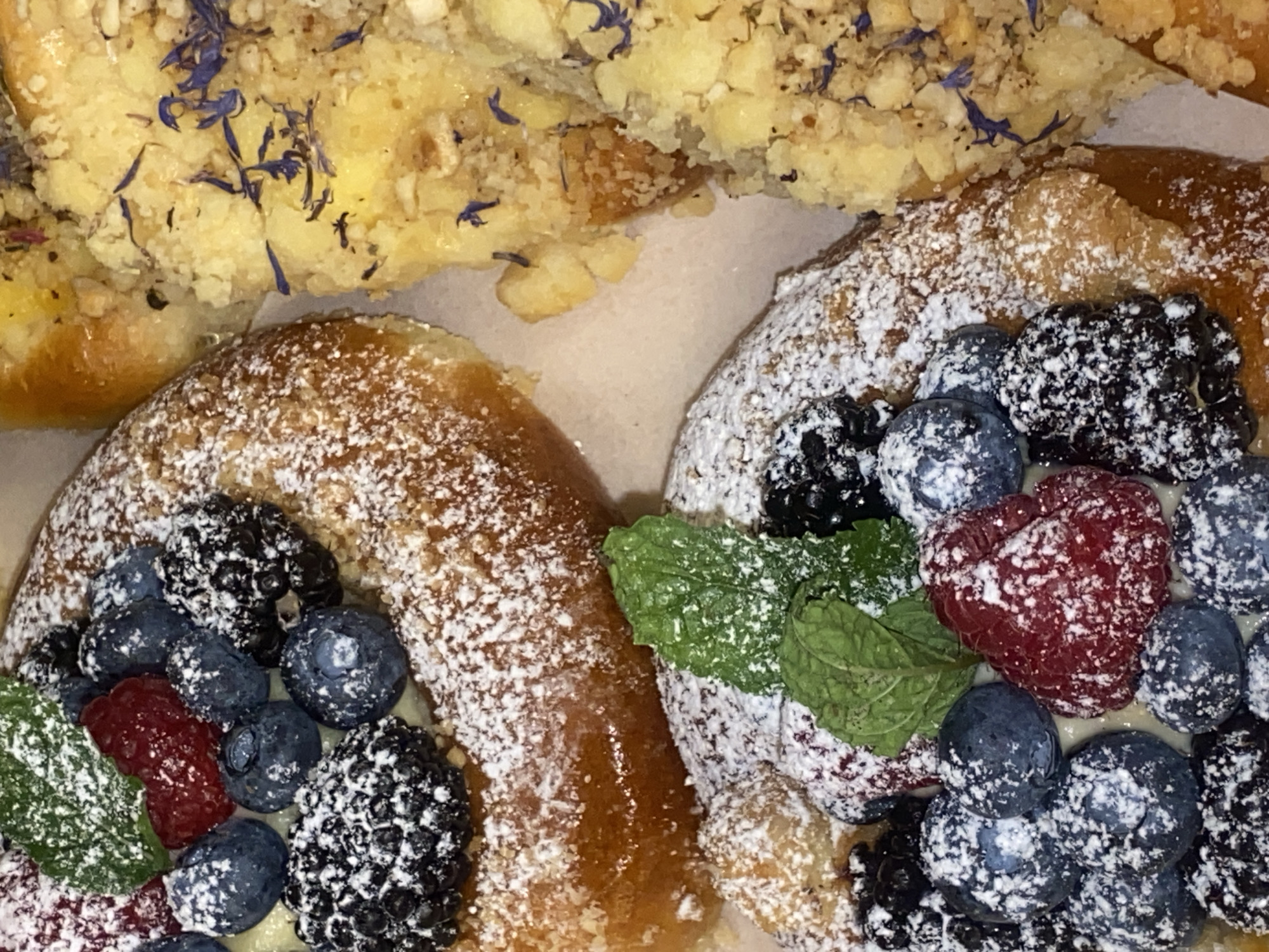 This close-up shot captures a variety of pastries dusted with powdered sugar, offering a visually appealing dessert spread. The pastries are arranged on a light-colored surface, hinting at a bakery or cafe setting.
The subjects are primarily food items. We see two distinct types of pastries. One is a larger, crumbly pastry topped with what appears to be streusel and delicate blue edible flowers, possibly cornflower petals. The other pastries are elongated, rolled buns or danishes, generously coated in powdered sugar. These are filled and topped with an assortment of fresh berries, including plump blueberries and vibrant raspberries, as well as dark blackberries. Fresh green mint leaves are artfully placed among the berries, adding a touch of color and freshness. A creamy filling is visible beneath the fruit on at least one of the rolled pastries.
The image does not depict any people or suggest a specific time of day or weather conditions, as the focus is entirely on the food. The setting is likely indoors, given the lighting and presentation, and could be a display case in a patisserie or a served plate at a cafe. The overall impression is one of deliciousness and a tempting treat. No visible text is present in the image. The context provided, Warsaw, Poland, adds a cultural layer to the culinary scene, suggesting these might be traditional Polish pastries or modern interpretations found in that city.