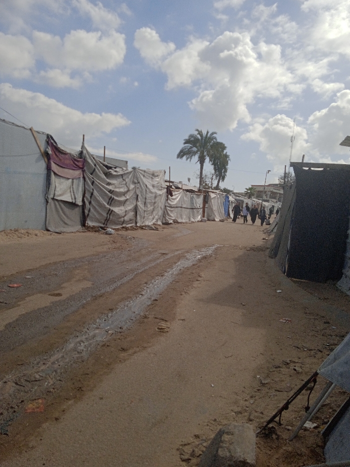 An outdoor, eye-level photograph depicts an unpaved dirt path in Khan Yunis, Palestinian Territory, flanked by makeshift shelters under a partly cloudy sky. The path, visible from foreground to mid-distance, features a central rut filled with standing water and mud. To the left, a continuous line of shelters constructed from grey and white tarpaulins, some with maroon fabric accents, extends into the distance. These structures utilize visible wooden poles and fabric for support. To the right, another line of similar shelters, including one prominent dark grey/black structure in the immediate foreground, defines the path's edge. In the mid-ground, a cluster of approximately six to eight individuals, primarily women dressed in dark clothing and head coverings, walk away from the camera along the right side of the path. Further in the mid-ground, several palm trees rise above the shelters. The background reveals additional, more permanent-looking buildings in the distance. The sky is blue with numerous scattered white cumulus clouds.