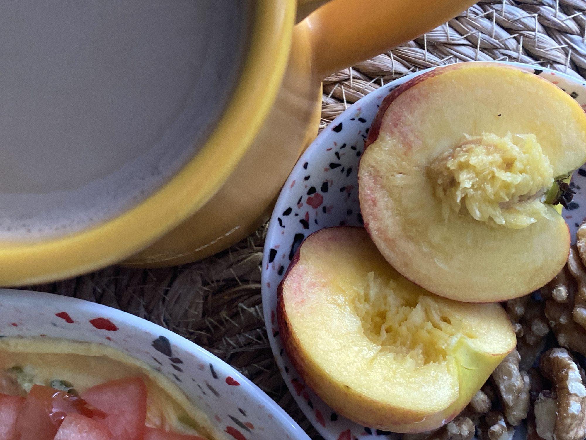 The image shows a close-up, top-down view of a breakfast setting, likely in Chaikovsky, Russia. In the foreground, two halves of a sliced peach sit on a small patterned plate. Next to the plate, a pile of walnuts is visible. Behind the peach plate, a portion of a yellow mug filled with a light-colored beverage, possibly coffee or milk, is present. To the left, another white plate with red and black speckles contains a dish with diced tomatoes and other ingredients, suggesting a meal in progress. The entire arrangement rests on a woven placemat. The lighting suggests natural daylight, and there are no individuals visible in the frame.