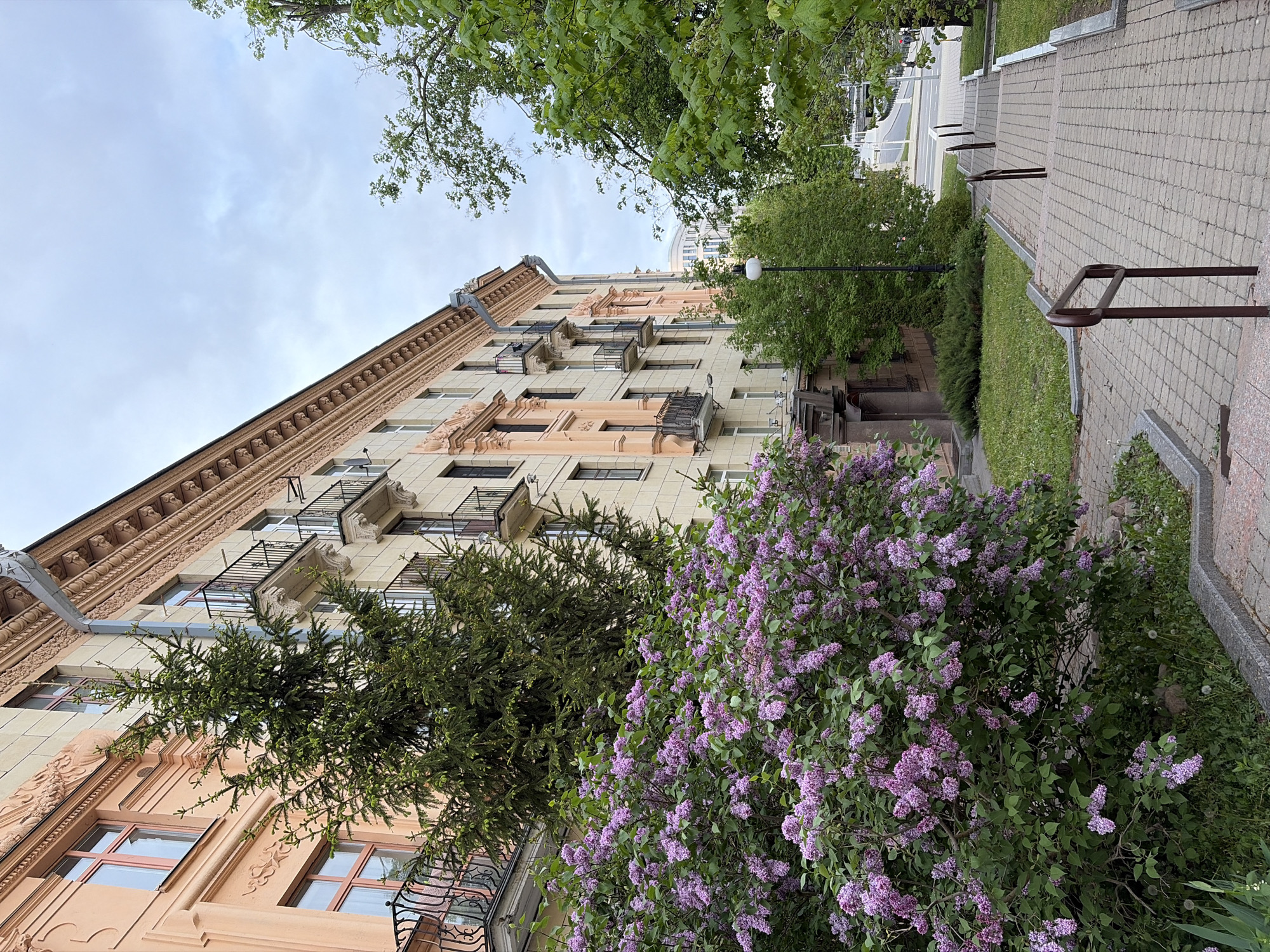 The image shows a building in Unknown, Unknown, taken from a low angle. The building is multi-story, with a mix of beige and salmon-colored facades, and features decorative moldings and small balconies. A drainpipe extends down the side. In the foreground, there is a large lilac bush in full bloom with purple flowers, partially obscuring the lower part of the building. To the left of the bush is a dark green coniferous tree. In the upper part of the image, there is a section of a paved walkway with handrails and green grassy areas on the sides. The sky is overcast.