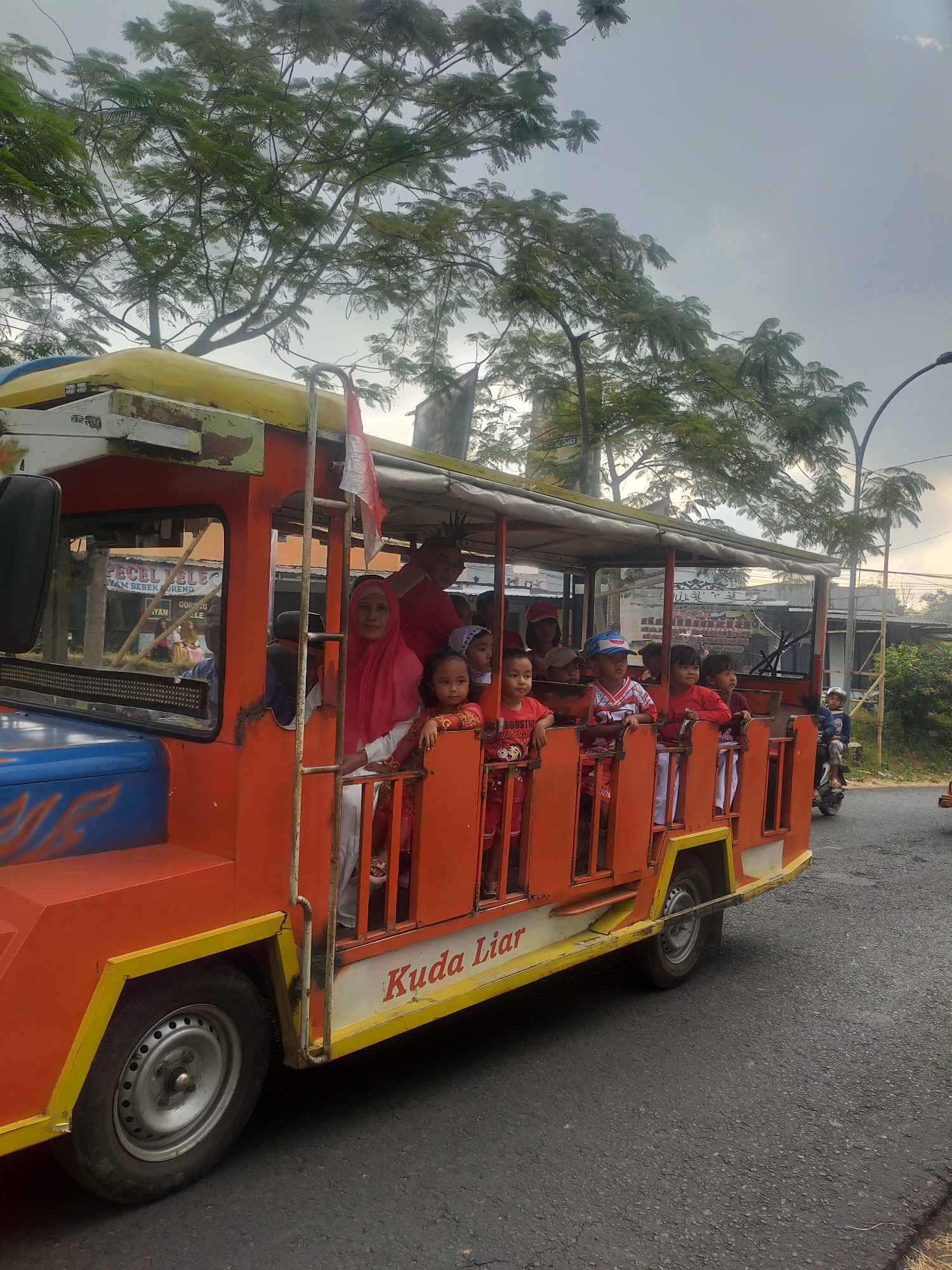 In Tuwel, Indonesia, a bright orange and yellow open-air vehicle, possibly a modified bus or truck, transports a group of passengers, predominantly children, along an asphalt road. The vehicle has a canvas roof and orange bars that act as protective barriers for the passengers. Most of the children are looking forward or to the side, with some smiling. A woman in a pink headscarf is visible in the front passenger area. In the background, trees with lush green foliage line the road, and a cloudy sky looms overhead. A street lamp is visible on the right side of the image. A motorcycle with a rider is also moving in the same direction as the vehicle. The word "Kuda Liar" is written in red on the side of the vehicle.