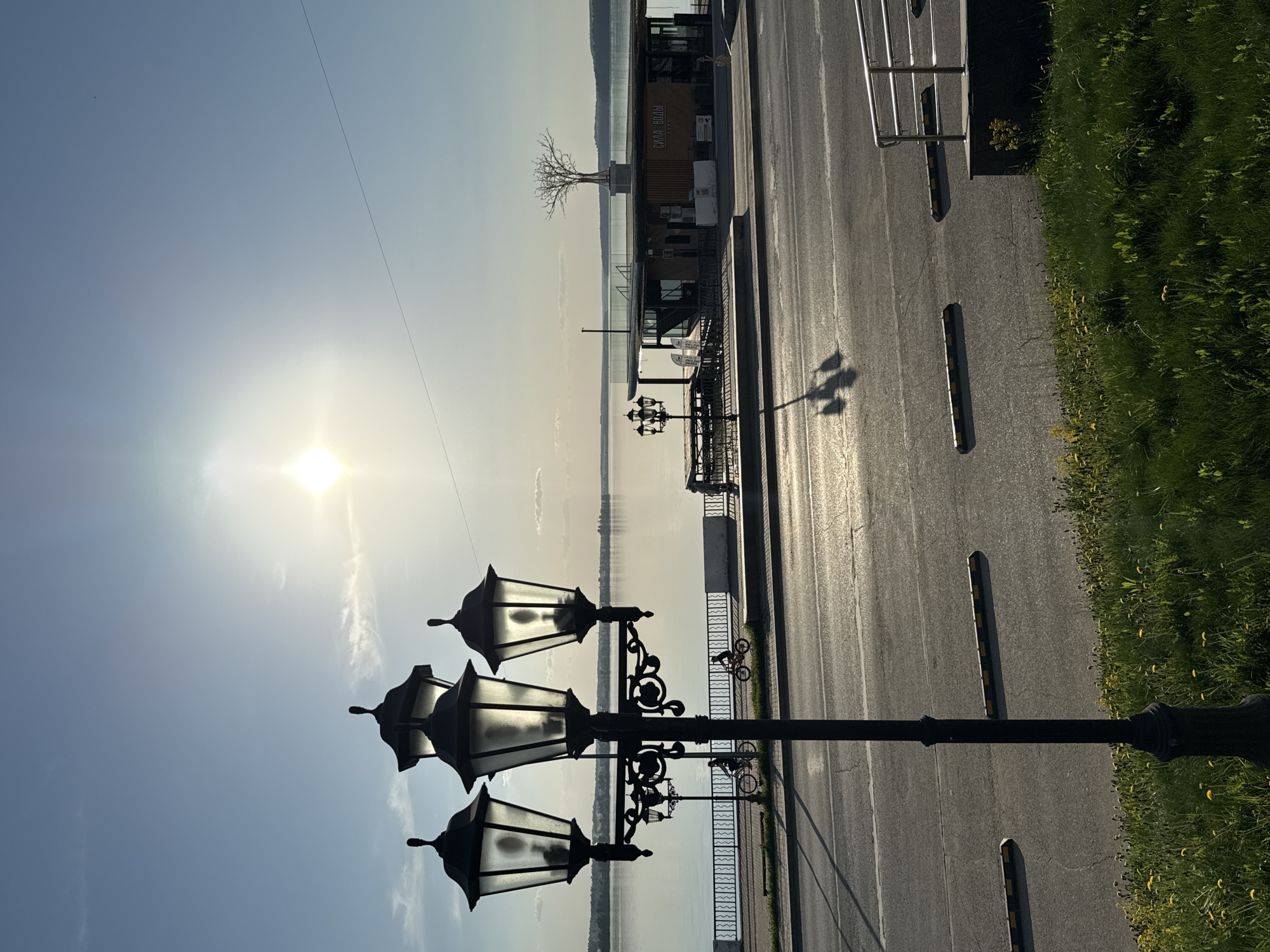 The image shows a street scene in Votkinsk, Russia, along a waterfront promenade. The sun shines brightly in the sky, reflecting off the water. In the foreground, a vintage lamppost with three lanterns stands prominently. To the right of the lamppost, a paved road runs parallel to the water's edge, with parking spots marked along its length. A patch of green grass lines the edge of the road. In the background, a modern building stands near the water, possibly a café or restaurant. A bare tree stands on top of the building. Decorative metal railings border the promenade near the water. The overall scene is bright and sunny, giving a sense of openness and tranquility.