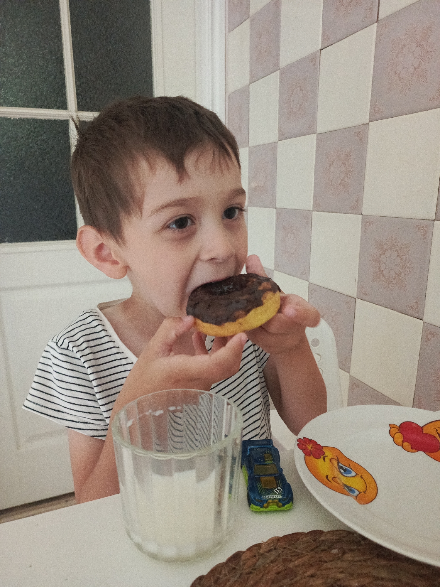 A young boy with brown hair is eating a donut with chocolate frosting. He is wearing a striped t-shirt. A glass of milk and a toy car are on the table in front of him. A plate with cartoon characters is also on the table. The background appears to be a tiled wall and a door. The scene is likely indoors, possibly in a home or a cafe, and it is not possible to determine the specific location within the city of Kurgan, Russia.