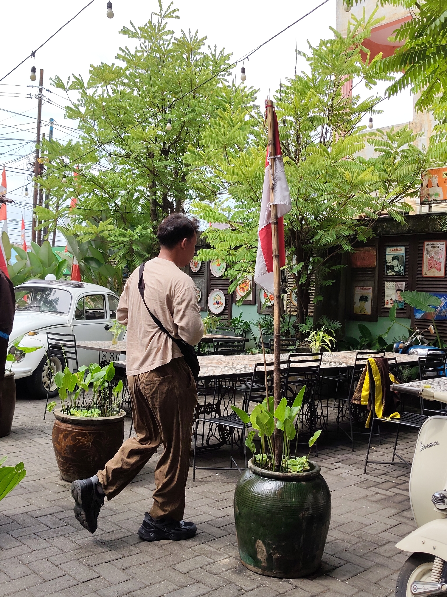 In the City of Medan, Indonesia, an outdoor seating area of a cafe or restaurant is depicted. A man wearing a tan t-shirt and brown pants walks across a paved patio. He is carrying a black bag slung across his chest. In the background, a white Volkswagen Beetle is parked. Several tables and chairs are set up for outdoor dining, and decorative elements such as string lights, potted plants, and framed pictures adorn the space. A red and white flag is visible, possibly indicating a national holiday. A light-colored Vespa scooter is parked on the right side of the frame. Lush green trees and foliage contribute to the outdoor ambiance.