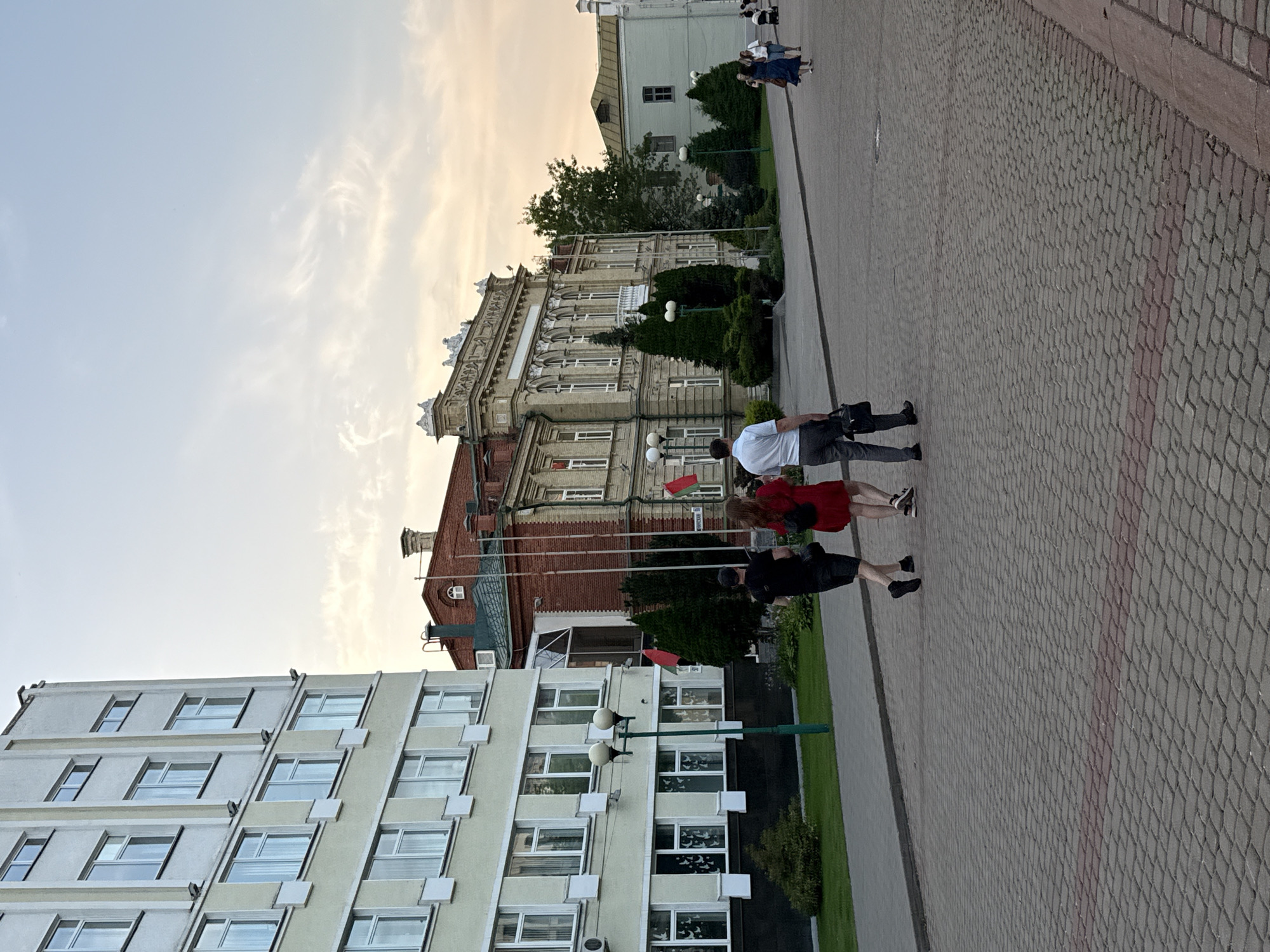 The image captures a view of a pedestrian area in front of administrative buildings in Mahilyow, Belarus. In the foreground, a paved walkway is visible, constructed with rectangular interlocking stones in a greyish hue, with a subtle red accent line running across it. Three individuals are walking away from the viewer on the walkway. The person on the left wears a black t-shirt and shorts. The middle person is dressed in a red dress, and the person on the right is in a white shirt and grey trousers, carrying a black briefcase.
Behind the pedestrians, a large, multi-story, pale yellow building with numerous white-framed windows dominates the left side of the frame. To the right of this building, a reddish-brown brick building with a green roof and ornate architectural details stands. Several red flags are displayed on poles in front of these buildings. Further to the right, a lighter green building with a light brown roof is visible, and in the distance, several more people are walking and some appear to be sitting. The sky above is a light blue with wispy clouds, suggesting either early morning or late afternoon light. The overall scene depicts a public space, likely a square or plaza, with architectural elements typical of an urban European setting.