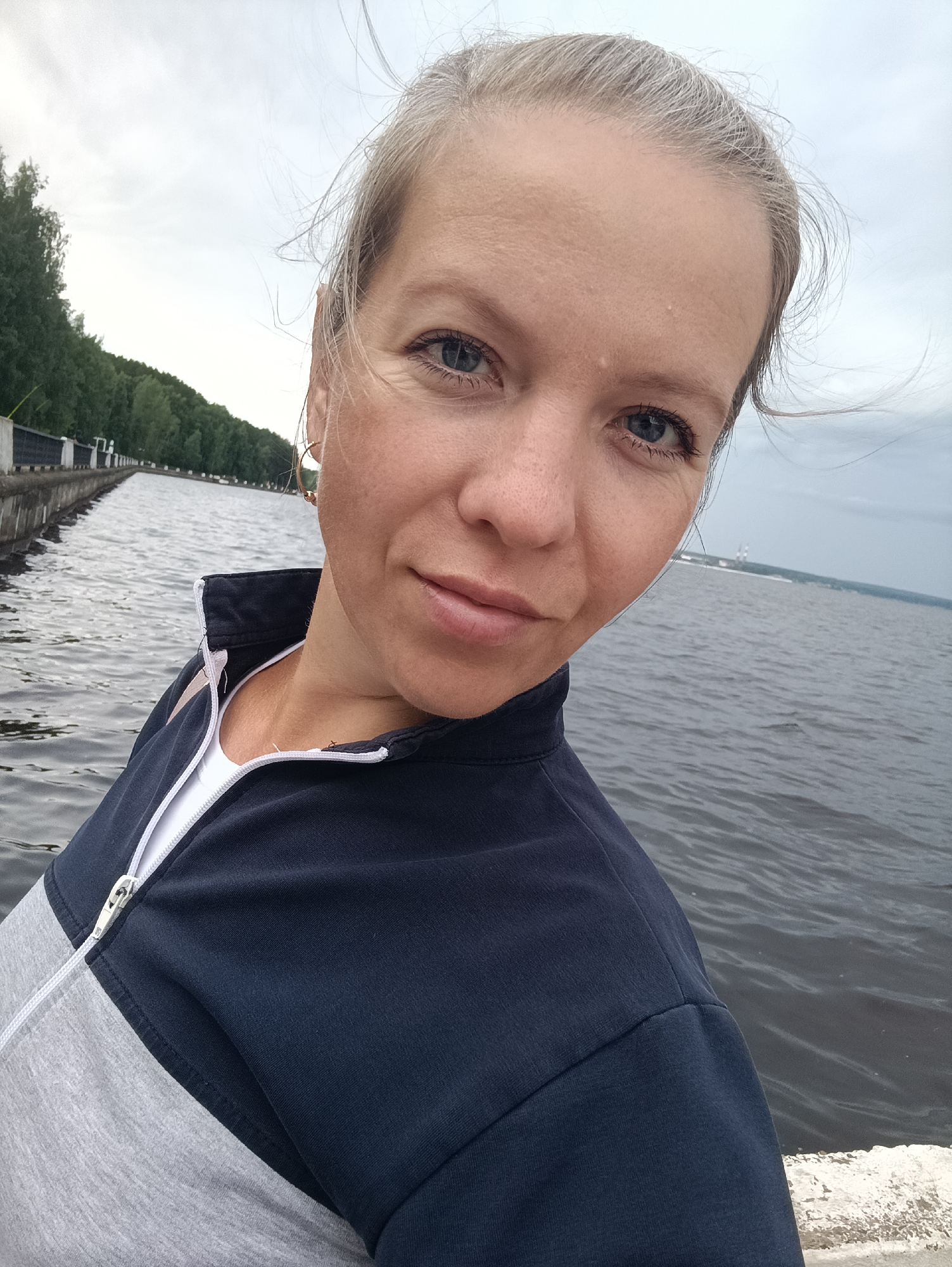 An outdoor, chest-up, selfie-style photograph shows a woman with fair skin and light blonde hair pulled back, looking towards the camera with a neutral expression. She is wearing a navy blue and grey zip-up jacket over a white t-shirt. Her left eye is in focus, and her right eye is partially visible. The background is a body of water with gentle ripples. On the left side of the frame, a concrete embankment with a metal railing runs along a treeline. In the distance, there is a hazy view of industrial structures. The lighting suggests an overcast day. The image is likely taken in the city of Chaikovsky, Russia, given the context of the embankment and industrial elements commonly found along waterways in the region.