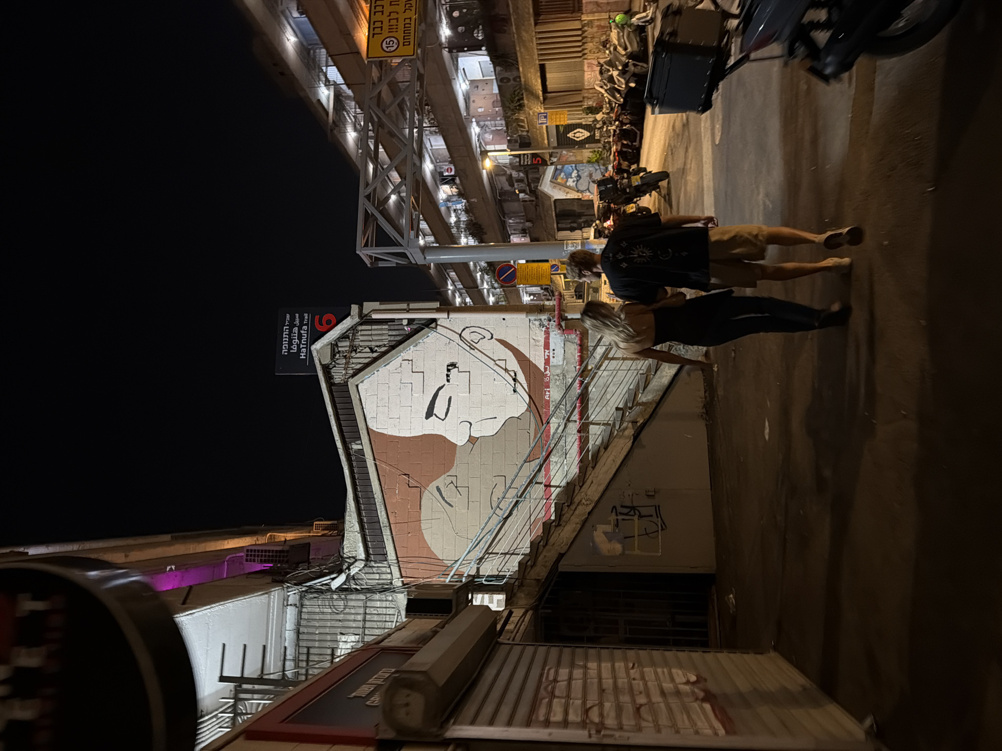 The image captures a nighttime street scene in Tel-Aviv, Israel, specifically near the "HaTnufa Trail" as indicated by a black sign with white Hebrew and English text ("שביל התנופה HaTnufa Trail") and the number "6" in red.
In the foreground, two individuals, a female and a male, walk away from the camera. The female, on the left, has blonde hair and wears a dark strapless top and dark pants. The male, on the right, wears a dark t-shirt with a light circular design on the back, light-colored shorts, and sneakers.
To the left, a large mural painted on a building wall depicts two stylized faces in profile, one with a lighter complexion and the other reddish-brown, positioned close together. Below the mural and adjacent to it, a concrete staircase with metal railings ascends. The lower section of the building to the left features a closed corrugated metal roll-down door. Above it, visible elements include utility wiring and what appears to be a purple light source along an architectural ledge.
Across the thoroughfare, to the right, a multi-story building with multiple illuminated balconies or open corridors is visible. Numerous parked motorcycles and scooters line the right side of the street. In the lower right corner, the rear of a motorcycle with a black storage box is partially visible.
Overhead, connecting structures between buildings, a grey metal truss-like framework spans the street. Several signs are present: a yellow sign on the right with black Hebrew text ("רכב כבד" - heavy vehicle) and a red circle containing the number "15", likely indicating a speed or weight restriction. A red circular prohibitory sign is visible on a pole in the mid-ground. Another pole features a small sign with a red "5" in a circle. The street surface is dark asphalt, reflecting some of the ambient light.