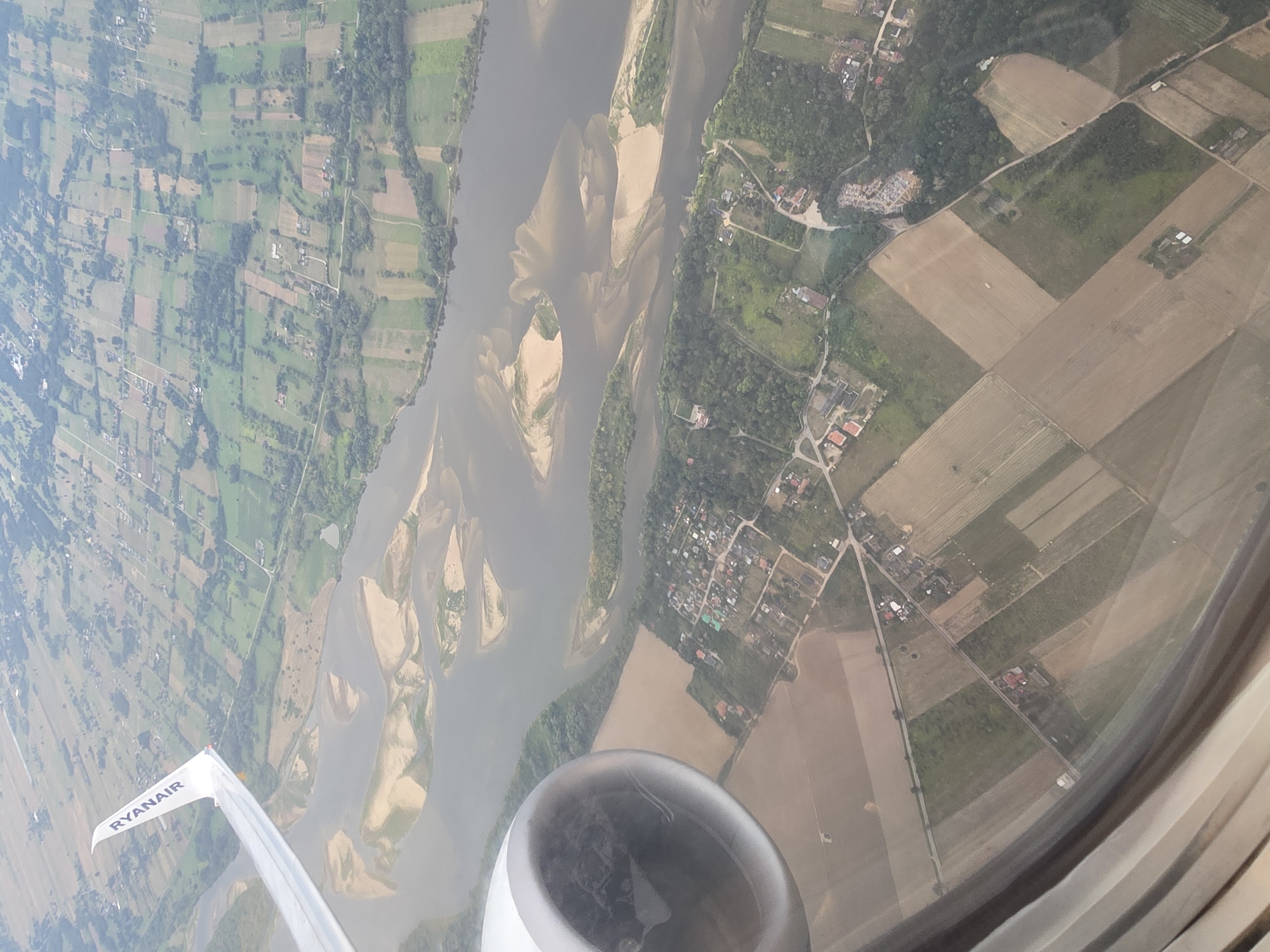 This is an aerial photograph taken from an airplane, likely in flight over a rural area. In the foreground, a portion of an airplane engine is visible on the left, and the airplane's wing, bearing the "RYANAIR" logo, extends into the upper left quadrant. The landscape below consists of a wide river with several sandbars and islands. The riverbanks are lined with trees and interspersed with agricultural fields, some appearing to be harvested, others with green crops. A small village or settlement is visible along one of the riverbanks, characterized by numerous small buildings with red roofs. The overall scene depicts a natural landscape with human habitation and agricultural activity from an elevated perspective. The image does not contain any identifiable features or context to suggest a location in the city of Kyustendil, Bulgaria.