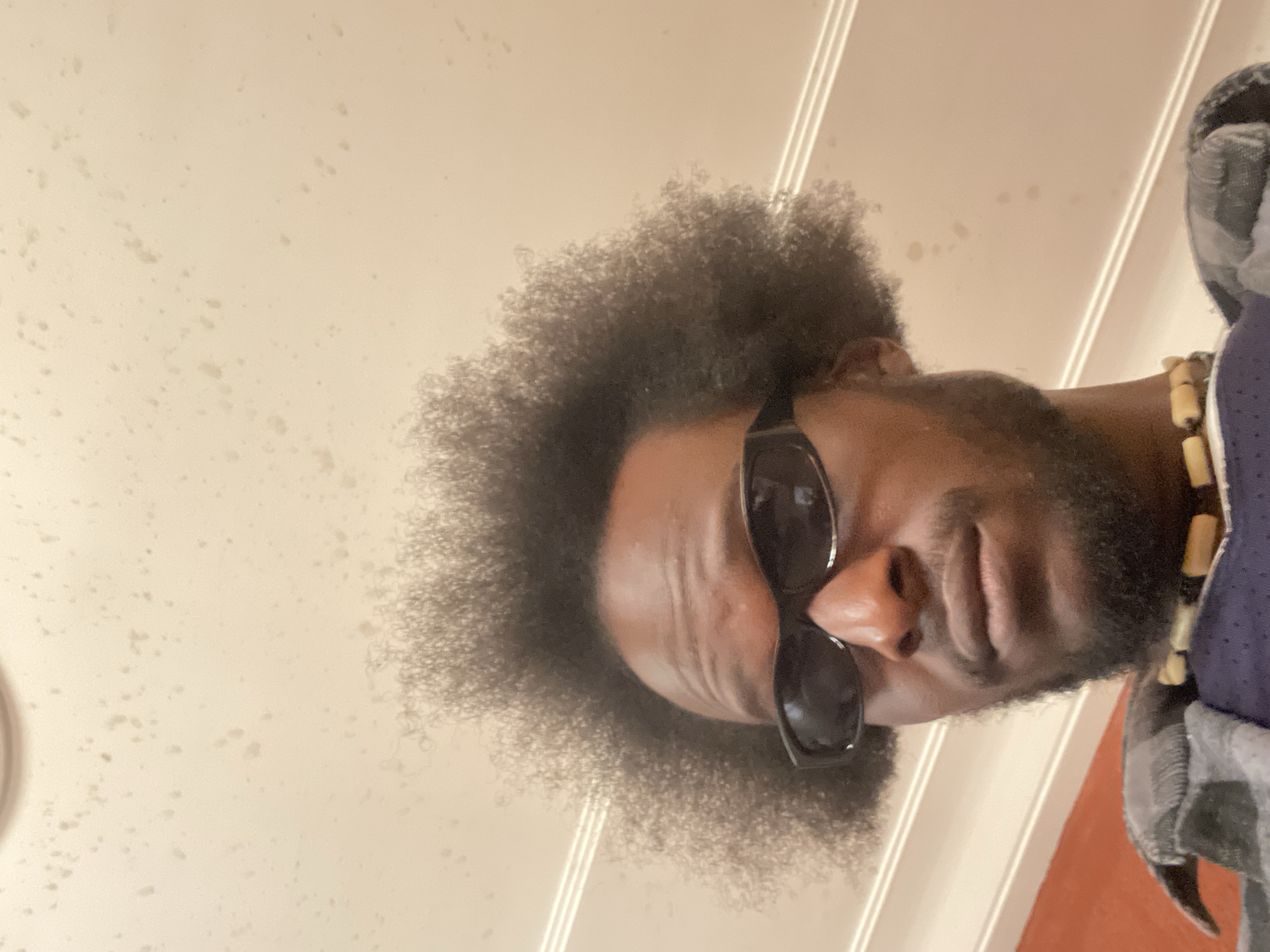 The image shows a close-up, low-angle shot of a Black man's face. He has an afro hairstyle, dark skin, and is wearing black sunglasses. He has a beard and a mustache. Around his neck, he wears a necklace with light-colored beads. He is wearing a plaid shirt over a dark blue garment. The background is a light-colored, textured ceiling with some scattered dark spots. There are horizontal lines visible on the wall or ceiling behind him. The image does not provide any information to suggest the location is the city of Jos, Nigeria.