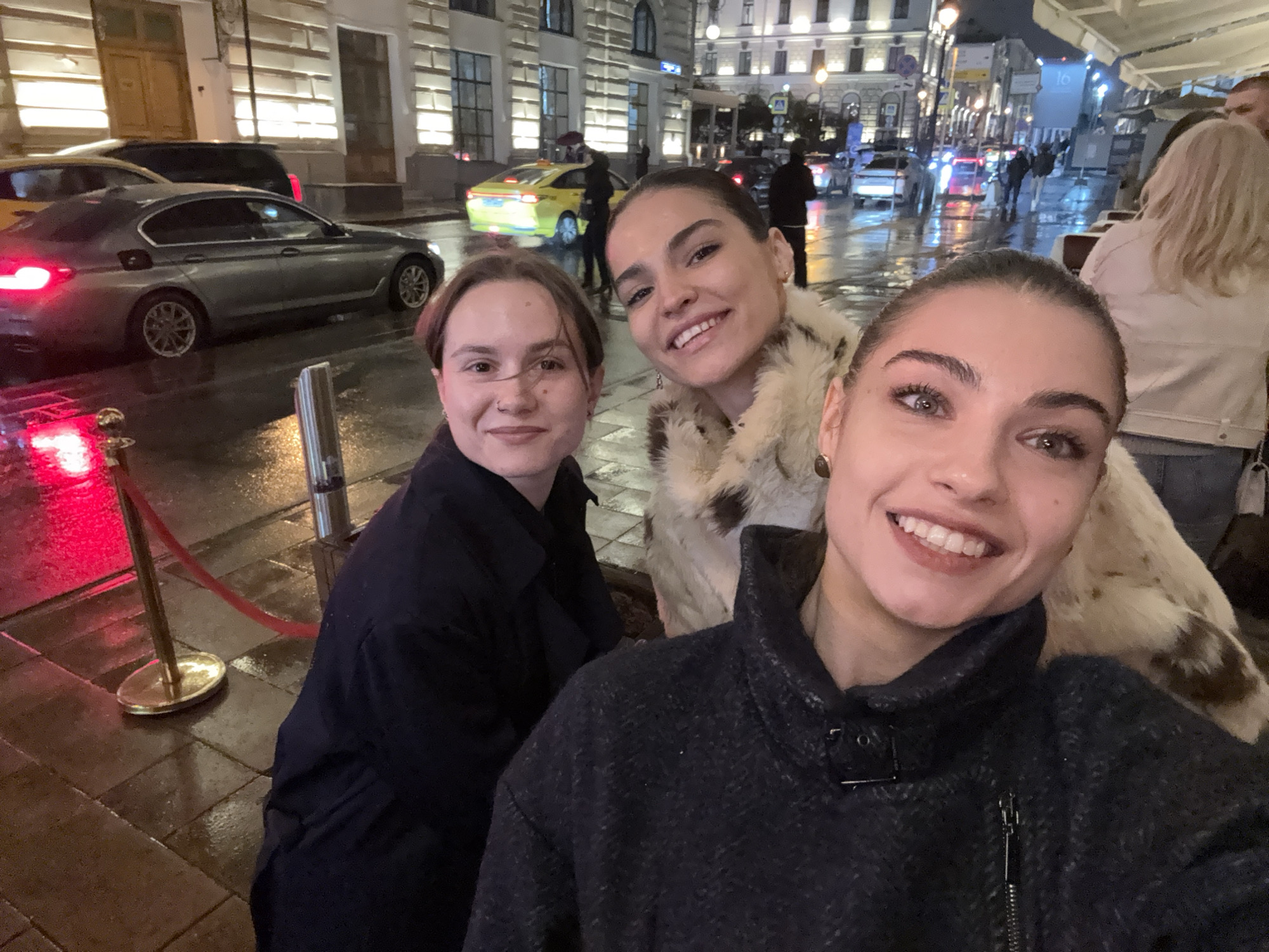 Three young women pose closely for a selfie. The woman on the right wears a dark gray jacket with a high collar and a zipper. The woman in the middle wears a light-colored fur coat. The woman on the left is wearing a black jacket or coat. All three are smiling.
Behind them, a city street scene unfolds at night. It appears to be raining or wet, as the pavement is shiny and reflects the city lights. There are several cars on the street, including a gray sedan, a black SUV, and a yellow taxi. There are other pedestrians visible, some walking with umbrellas. Buildings with illuminated windows line the street, and streetlights are visible. A red velvet rope on a stand is visible near the bottom left of the image.