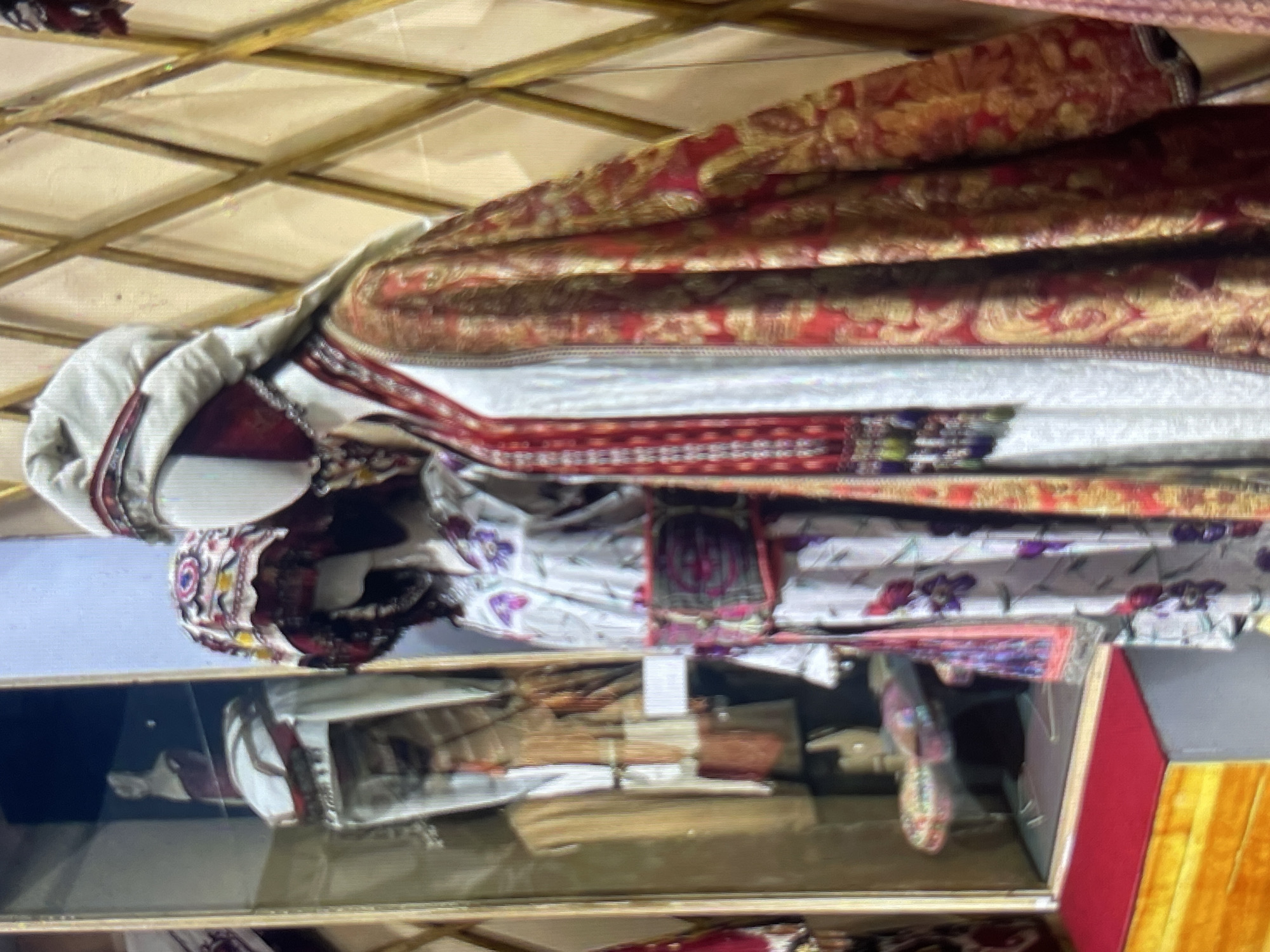 The image shows a display of traditional Kyrgyz clothing, likely in a museum in Bishkek City, Kyrgyzstan. Two mannequins are dressed in ornate garments. The mannequin on the right wears a white robe with a richly embroidered red and gold outer garment, a matching headpiece, and a decorative sash. The mannequin on the left is adorned in a white robe with a floral pattern, a colorful sash, and a patterned headdress. In the background, a display case contains other traditional clothing items, and the wall behind the mannequins is constructed from wooden lattice work. The overall impression is of a cultural exhibit showcasing traditional Kyrgyz attire.