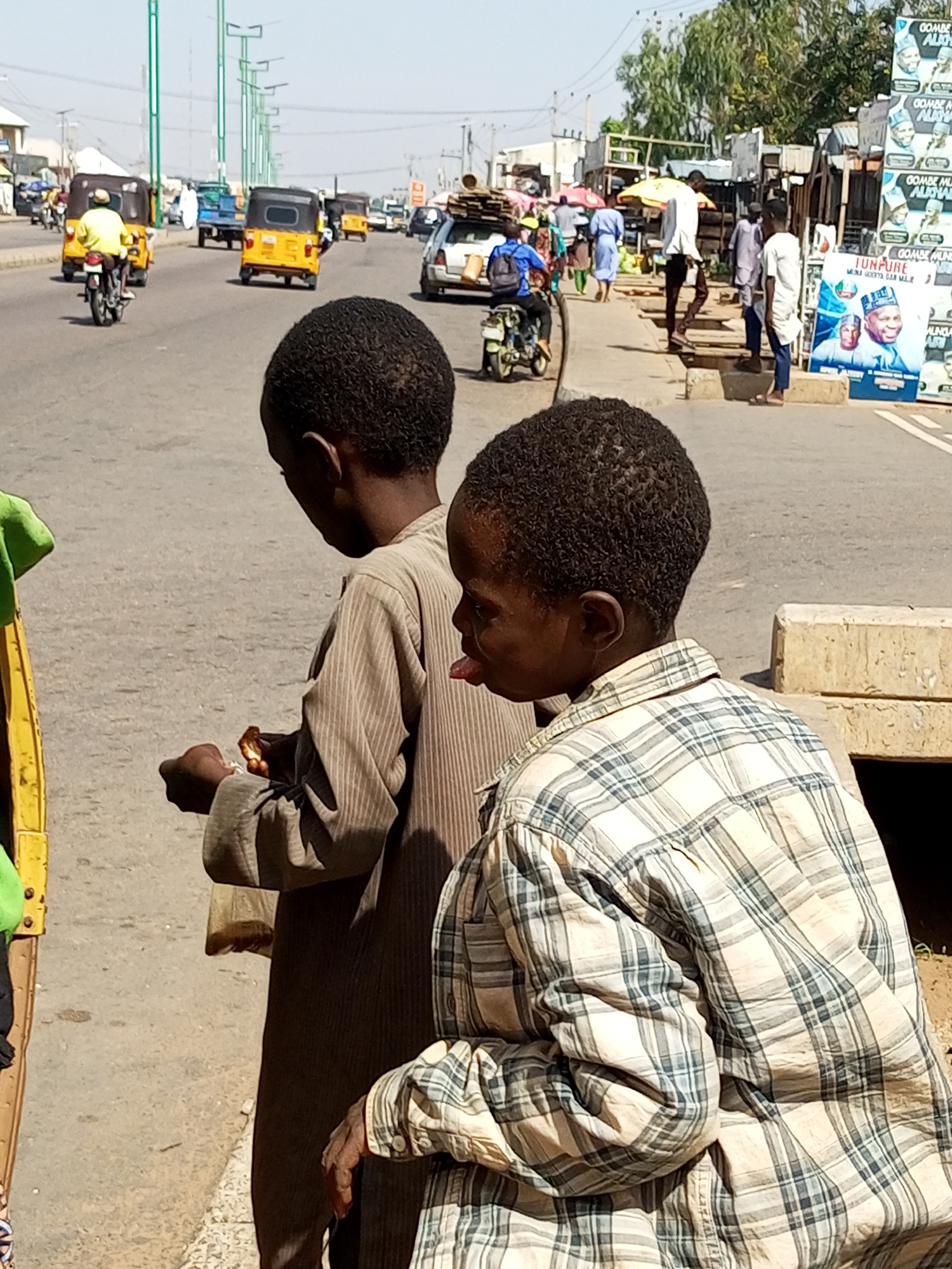 In Gombe, Nigeria, two young boys stand by a roadside. The boy on the left, wearing a brown striped tunic, holds a piece of food in his right hand. The boy on the right, wearing a plaid shirt, sticks his tongue out. Behind them, a busy street is filled with vehicles, including yellow auto rickshaws and motorbikes. People walk along the sidewalk, some carrying goods. Large posters are visible on the right side of the street, suggesting a commercial or political area. The environment appears to be an urban setting with clear skies.