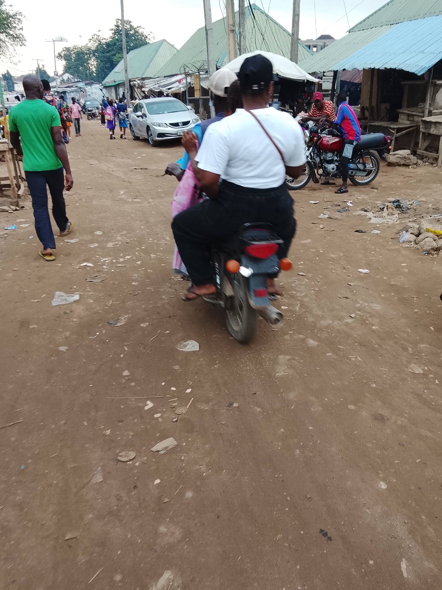 The image depicts a busy street scene in Abuja, Nigeria, with a dirt road and several people. In the foreground, a person is riding a motorcycle, with another person seated behind them, partially obscured. The rider is wearing a white t-shirt and black pants, while the passenger is wearing a pink fabric. The motorcycle is red and black.
In the background, several other people are walking along the road. One person in a green t-shirt and blue jeans is walking away from the camera. A silver car is parked on the side of the road. Various stalls or shops with corrugated metal roofs line the street, suggesting a market or commercial area. The environment appears dusty and unpaved, typical of some areas in Abuja. There is debris scattered on the ground, including plastic waste. The overall impression is a vibrant, albeit somewhat unkempt, urban street life.