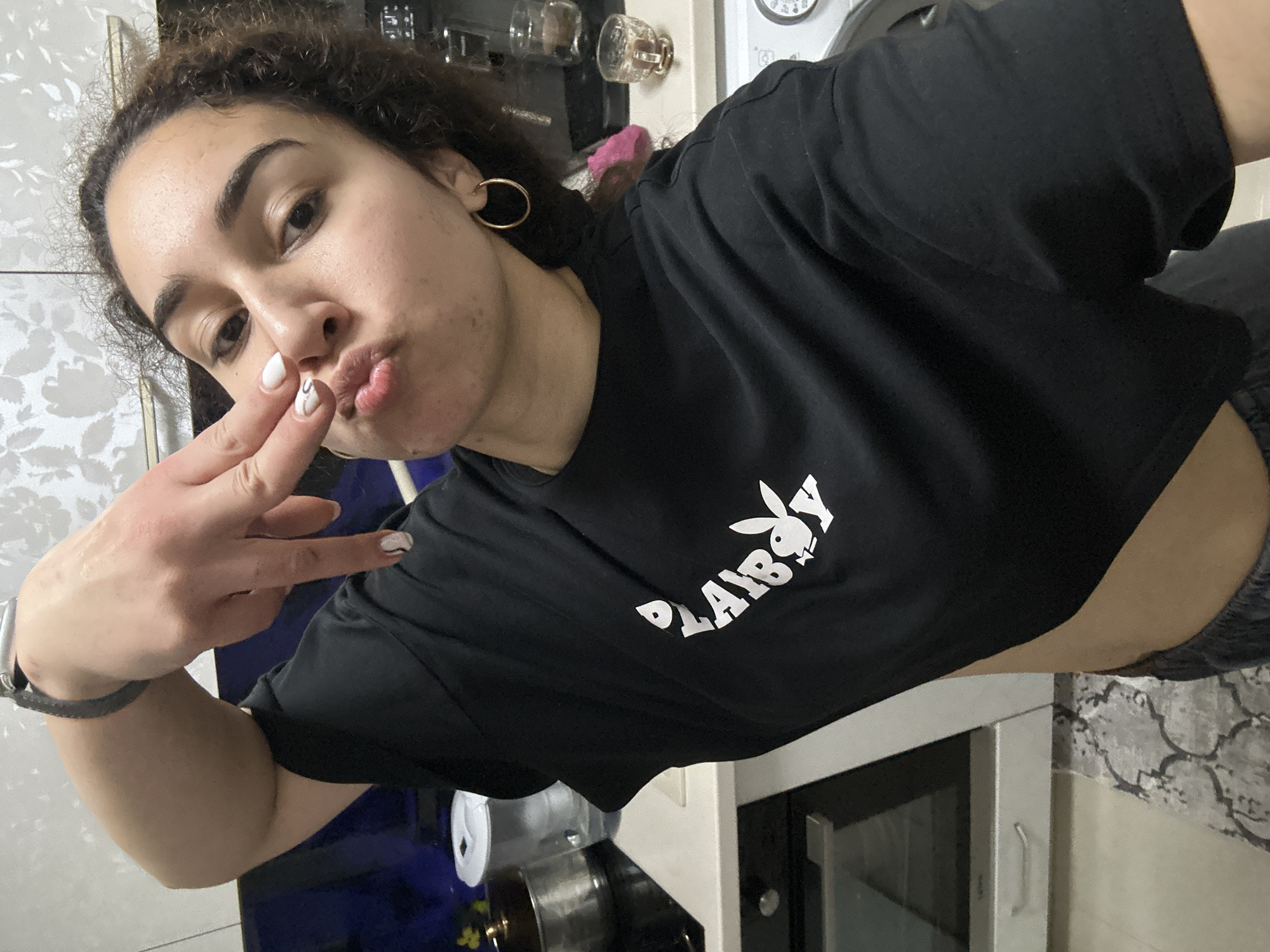 A young woman with curly brown hair and light skin is taking a selfie in what appears to be a kitchen. She is wearing a black t-shirt with the Playboy logo in white. She is making a kissing face and holding up two fingers, showing her nails that are painted white with a black design. She has a gold hoop earring in her left ear and a dark-colored watch on her left wrist. The background includes kitchen cabinets, appliances, and cookware. There is no information available to determine a location in Burgas, Bulgaria.