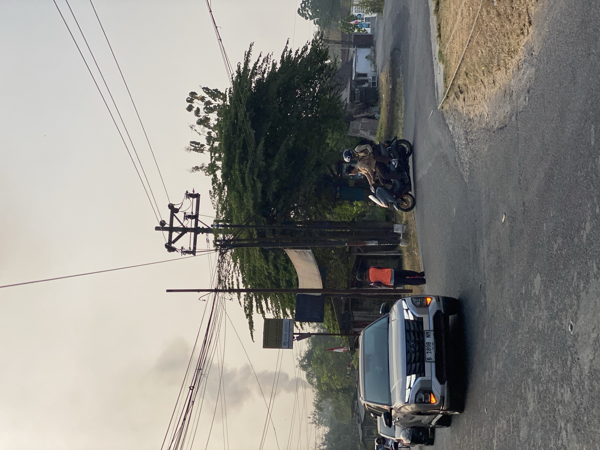 In the city of Kasihan, Indonesia, on a clear day with an overcast sky, a street scene unfolds. A modern silver SUV, bearing the license plate "B 1898 NM," is positioned in the foreground, facing right and slightly towards the viewer. To the left of the SUV, a portion of another vehicle is visible. On the right side of the road, a dark-colored scooter carries two individuals dressed in tan uniforms and helmets. One person is driving, and the other is a passenger. Further down the road, a person wearing an orange vest walks away from the camera. In the background, a large, dense green tree dominates the landscape, with buildings and more trees visible beyond it. A prominent utility pole with numerous wires stretches across the upper portion of the image. Wisps of smoke are rising in the upper left quadrant, indicating a potential fire or industrial activity. A sign for "BANK BANTUL" is affixed to a pole behind the SUV.
