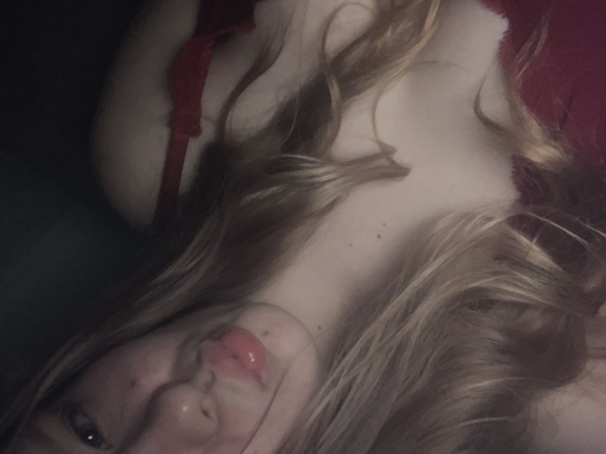 A close-up, dimly lit image shows a person, likely female, from a low, angled perspective. The subject has long, wavy blonde or light brown hair that falls across their chest and face. They are wearing a red garment, possibly a bra or top, with one strap visible over the shoulder.
The face is partially visible towards the bottom left of the frame. One eye is open and looks towards the viewer. The nose and mouth are also visible, with the lips slightly parted and appearing glossy or made up with a reddish tint. Small moles or freckles are scattered on the visible skin of the chest and neck. The overall lighting is low, suggesting an indoor setting, possibly in the evening or night. The pose appears relaxed, as if the person is lying down and taking a selfie. There is no visible text in the image. The implied location context is Saint Petersburg, Russia.