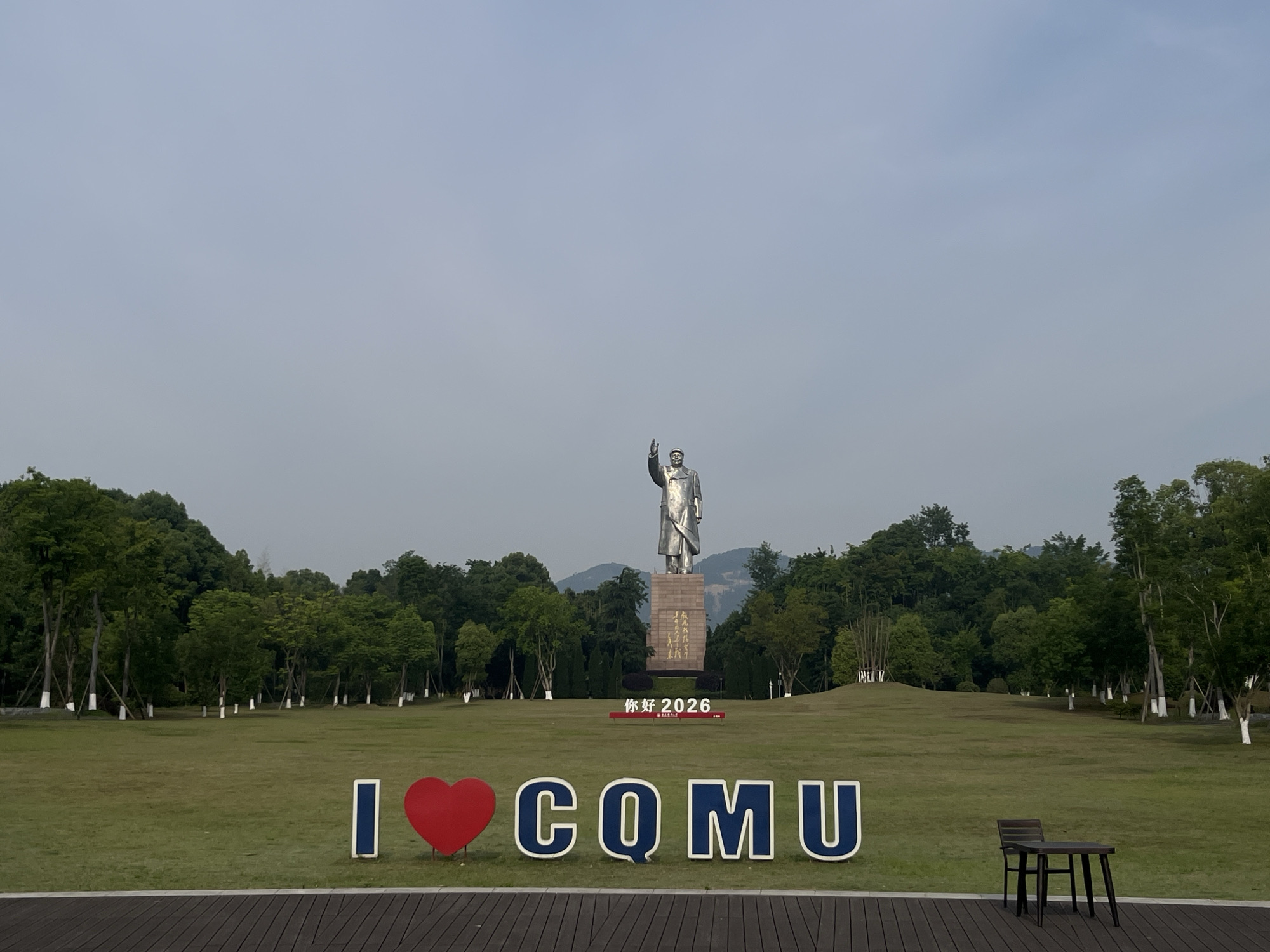 The photograph captures a park scene in the Chongqing High-tech Zone, China. Dominating the middle ground is a statue of Mao Zedong atop a stone pedestal. A sign beneath the statue reads "你好 2026," which translates to "Hello 2026" in English. In the foreground, large letters spelling "I ❤️ CQMU" are placed, where the heart symbol replaces the word "love." A dark wooden table and two chairs sit to the right of the letters. The setting includes a broad grassy field, flanked by dense trees and shrubs. A hint of mountainous terrain is visible in the background behind the trees, under a hazy sky. The foreground features a dark wooden platform or deck.