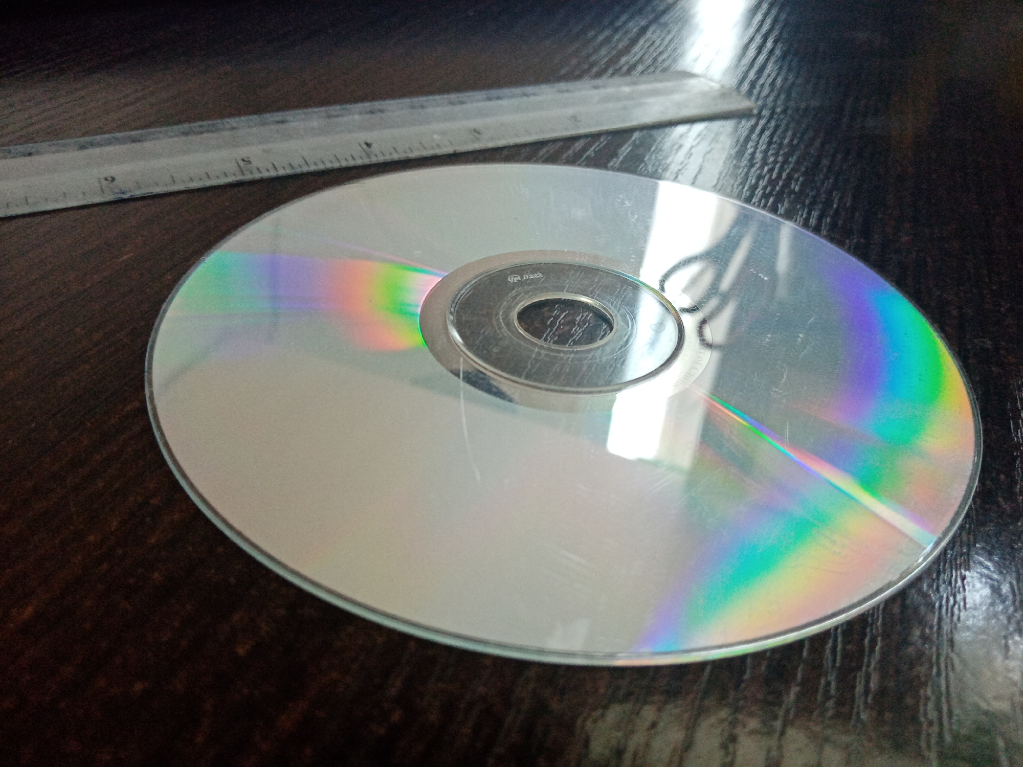 The image shows a compact disc and a ruler on a dark, textured surface. The CD exhibits a rainbow iridescence pattern. The ruler is metallic and marked with measurements. There is no discernible indication of the location being Elektrostal, Russia. There are no people visible.