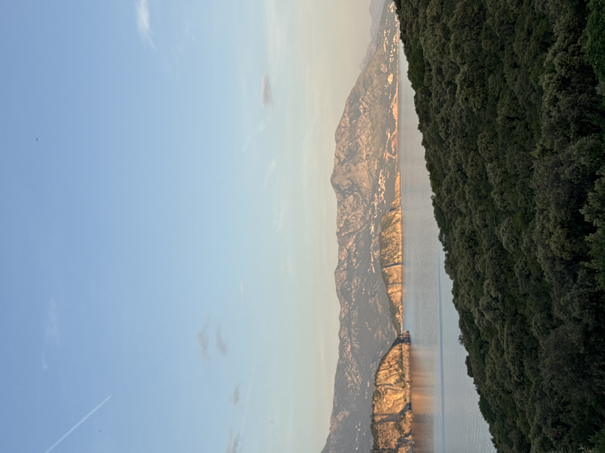 The image shows a landscape featuring a mountain range, a body of water, a forested area, and a blue sky with wispy clouds. The location is Dubraljevina, Montenegro. The mountain range is rugged and appears rocky, with some signs of vegetation. The body of water seems calm and reflects light. The forest covers a significant portion of the right side of the image, exhibiting dense, dark green foliage. The sky is clear with a gradient of light blue to pale yellow, suggesting either sunrise or sunset. A contrail is visible in the upper left part of the sky.
