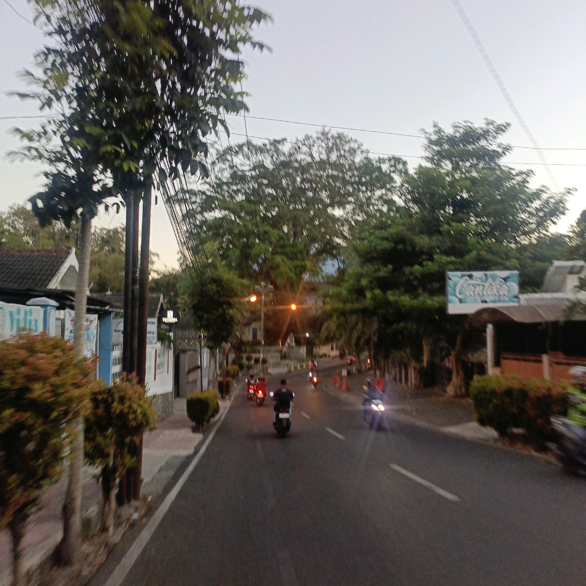 The image captures a street scene in Salatiga, Indonesia, likely during the early evening or late afternoon. A paved road occupies the foreground and midground, with a white line marking the edge of the lane. Several motorcycles are present, with some riders visible. One motorcycle in the foreground has its headlights illuminated, and another in the midground has a rider wearing a dark shirt and helmet. Further down the road, more motorcycles are seen, some with riders.
On the left side of the road, a sidewalk runs alongside a fence and a building with a blue and white facade. Trees and bushes are planted along the sidewalk, some of which are trimmed into hedges. Power lines are visible crisscrossing the sky above the trees. On the right side of the road, there is a fence, more foliage, and a building with a prominent sign that appears to say "Candika" in stylized lettering. Traffic cones are also visible on the right side of the road. The overall impression is of a moderately busy street in a residential or commercial area.
