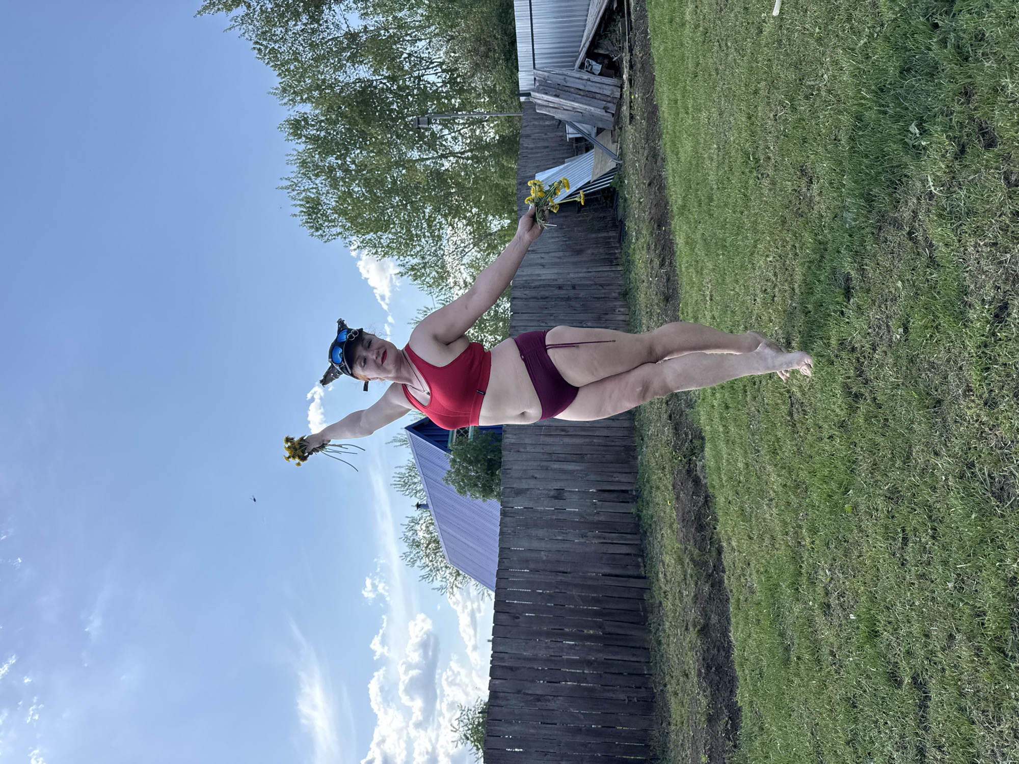 A woman is horizontally oriented in an outdoor setting. She is wearing a red sports bra, maroon shorts, and what appears to be a head-mounted device with goggles. She extends her arms outward, holding yellow flowers in her left hand. The background includes a green lawn, trees, a wooden fence, a structure with a metallic roof, and a partially clouded blue sky. The location is Chaikovsky, Russia.