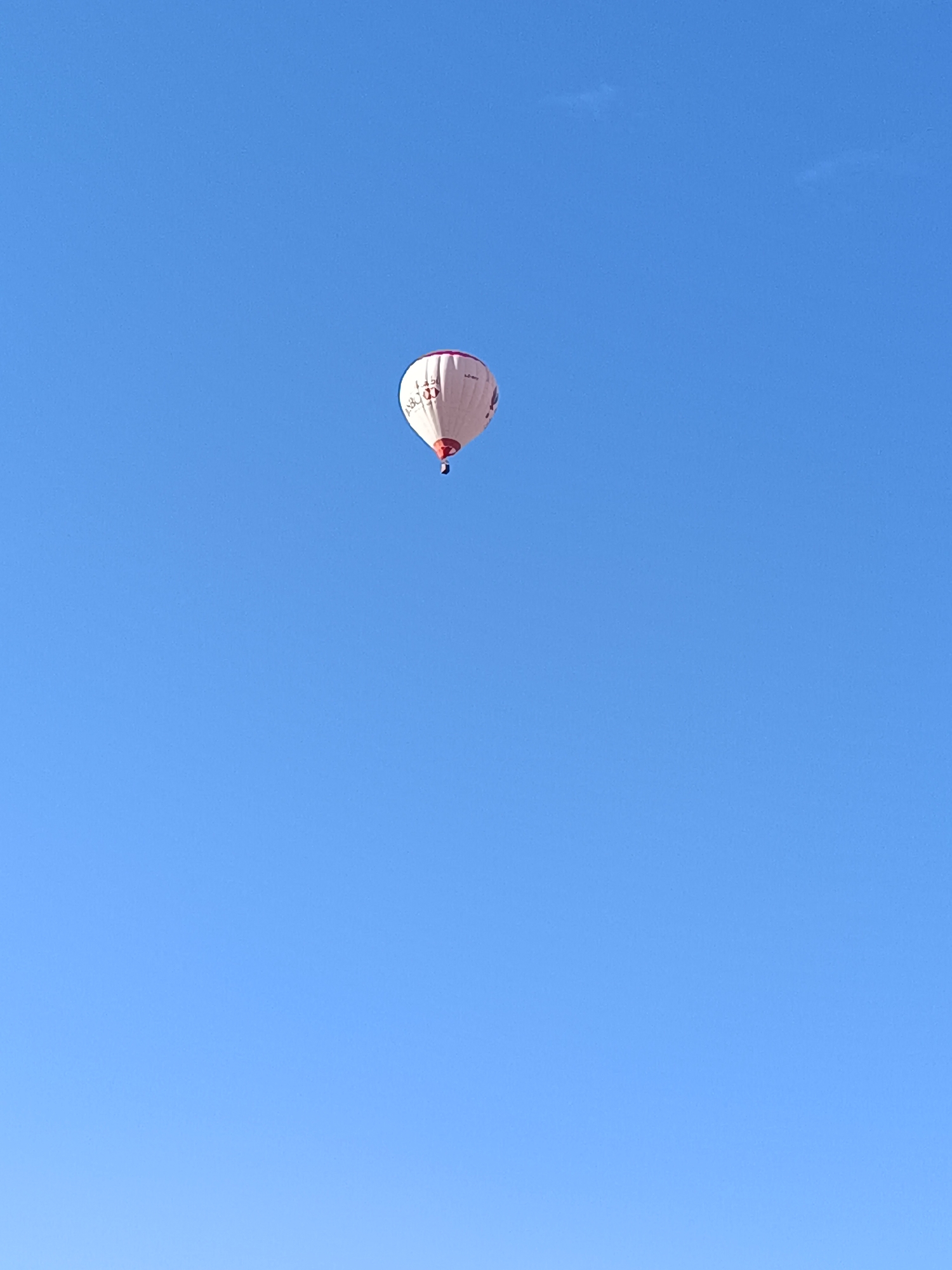 A single hot air balloon floats peacefully against a vast, clear blue sky. The balloon is white with a prominent red band around its lower portion and red lettering on its main body. The most discernible text on the balloon is the "HSBC" logo, rendered in red capital letters. Above this, smaller red text, possibly a website or slogan, is partially visible, and to the right, a registration code starting with "G-" can be faintly seen.
The setting is an open sky, completely devoid of clouds, indicating excellent weather conditions. The bright, even lighting suggests it is daytime, likely morning or early afternoon. The balloon is relatively small in the frame, emphasizing the immense expanse of the sky. The scene is tranquil and serene, capturing the quiet ascent or flight of the balloon. The location context for this image is Vasil Drumev, Bulgaria.