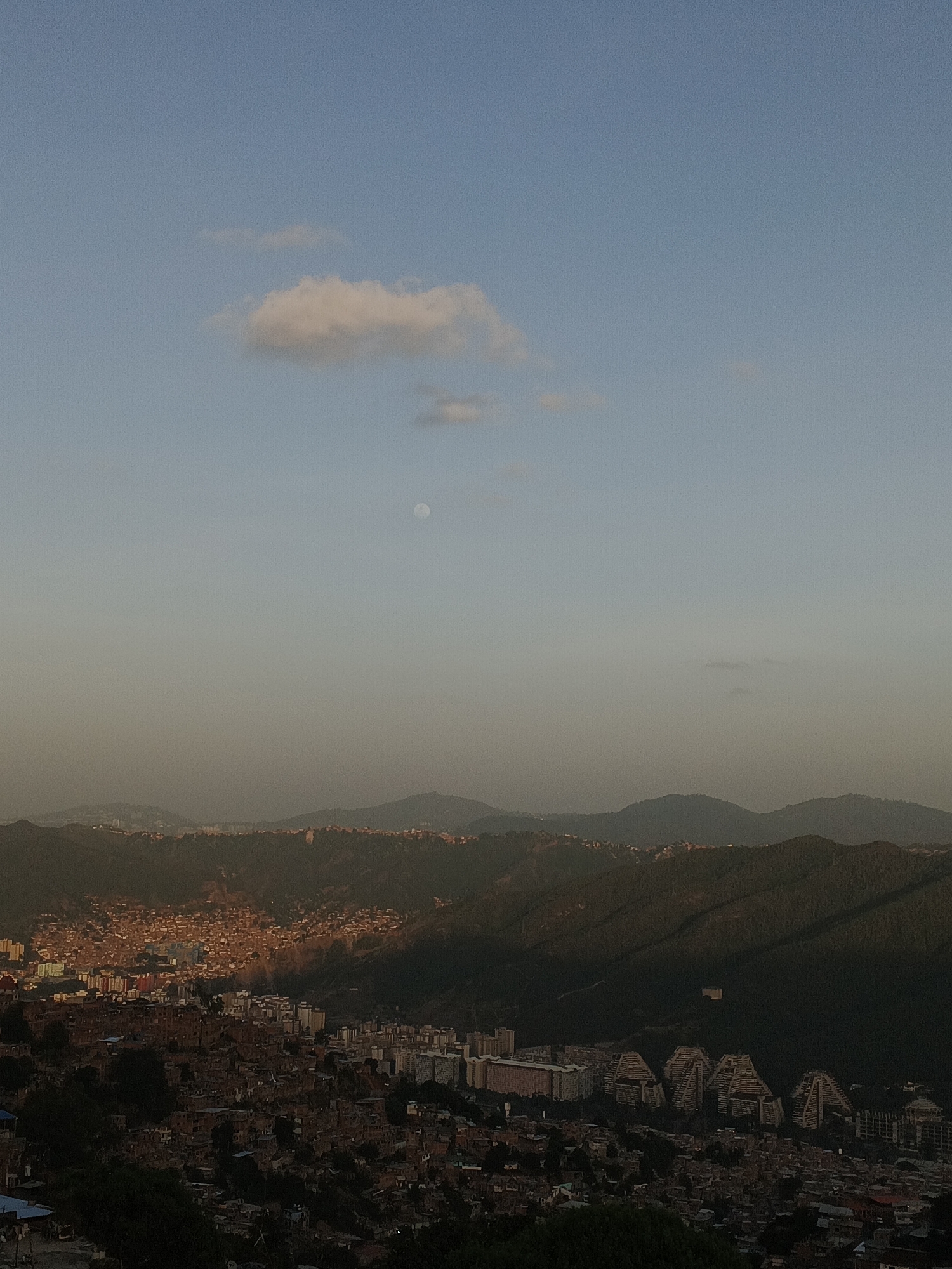 The image shows a high-angle, daytime view of Caracas, Venezuela, looking towards the horizon with a visible sky featuring a single cloud and a faint moon. The foreground and mid-ground are dominated by residential areas, varying in density and architectural style from tightly packed informal settlements on the left to higher-density apartment buildings. The hills surrounding the city are visible behind the residential area with a clear color shift of the vegetation on them. In the background, distant mountains are silhouetted against the sky. There are no people visible in the image.