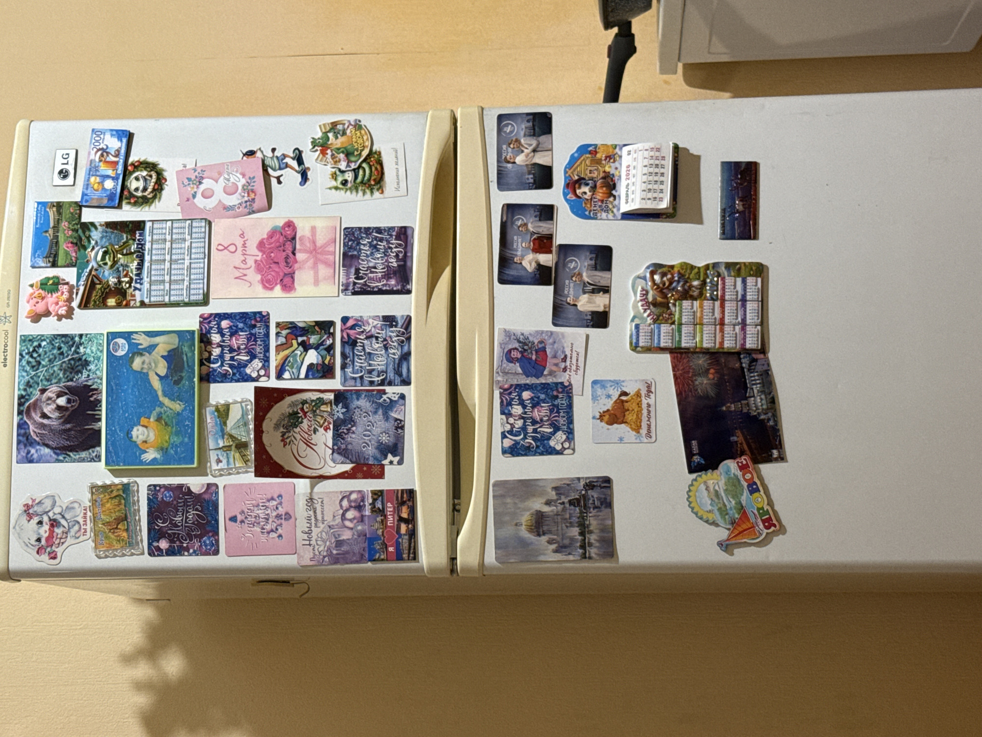 The image shows the side of a white refrigerator covered in various magnets. These magnets include a picture of a bear, people swimming, a pig, buildings, a calendar, and holiday-themed decorations. The refrigerator is situated in a room with a light yellow wall. The lower portion of the refrigerator has a silver handle, and part of a black saucepan handle is visible on the right side of the frame, indicating a kitchen environment. There are no people in the image.
The provided information about the location in the city of Kholmsk, Russia is not verifiable from the visual content of the image itself. There are no landmarks or context clues within the photograph that directly indicate this specific location.