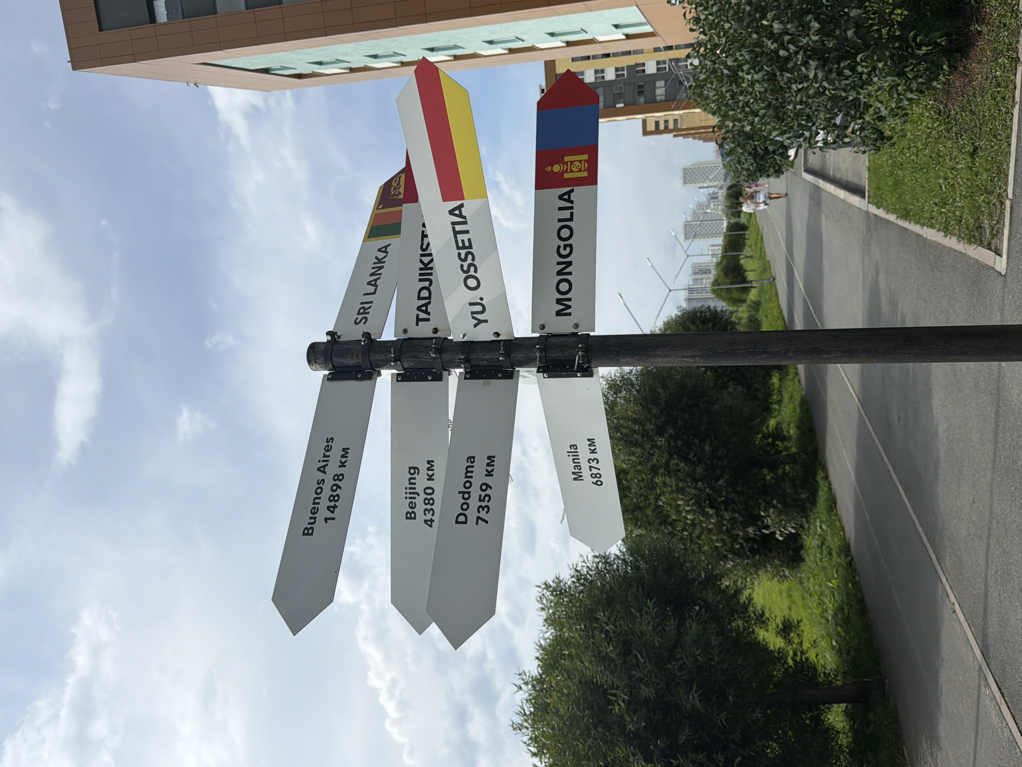 A directional signpost stands on a paved path in a city park. The signpost has multiple white directional arrows, each pointing to a different city and listing its distance in kilometers. Some arrows also feature flags of the respective countries. The arrow closest to the top points to "Buenos Aires 14898 KM". Below that, an arrow points to "Beijing 4380 KM". Further down, an arrow indicates "Dodoma 7359 KM", and another points to "Manila 6873 KM". On the right side of the signpost, arrows point to "Sri Lanka", "Tadjikistan", "Yu. Ossetia", and "Mongolia", each with a corresponding flag. In the background, there are tall residential buildings, trees, and streetlights lining the path. Two people are walking away from the camera on the path. The sky is partly cloudy. The signpost is located in Yekaterinburg, Russia, specifically in the "Park of the Great Love" (Парк Влюблённых) which features such a directional sign.