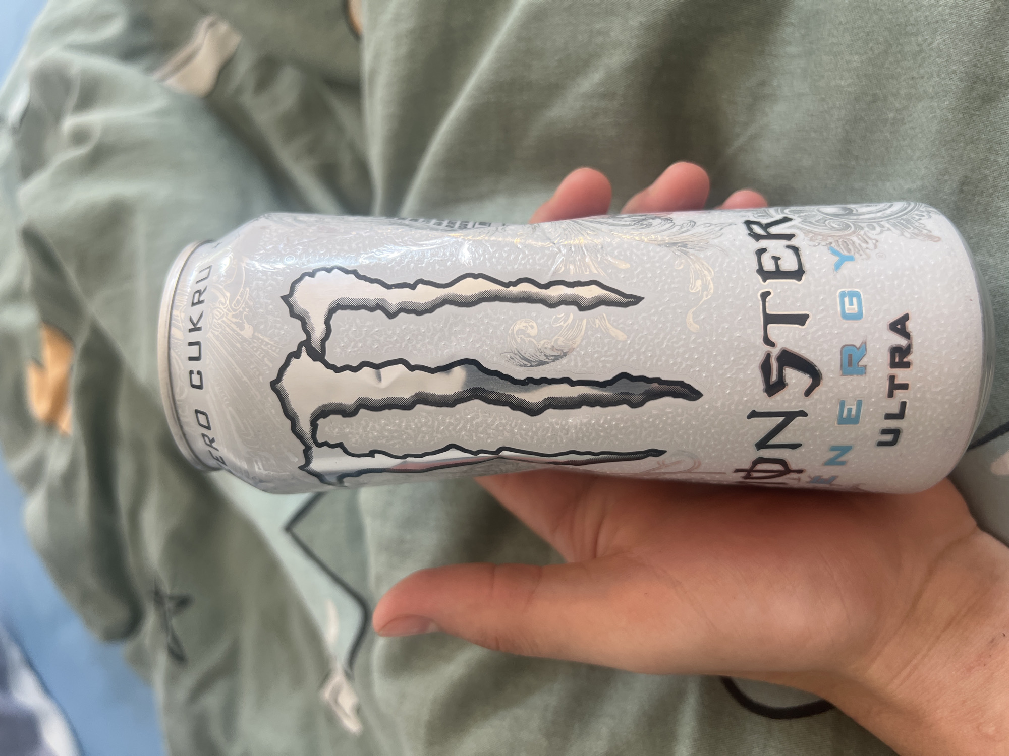 A hand is holding a white can of Monster Energy Ultra drink. The can has the Monster Energy logo and "ZERO CUKRU" written on it. The background is a green-blue patterned blanket. The scene appears to be indoors. The location of Szczebrzeszyn, Poland is not depicted or suggested by the image.