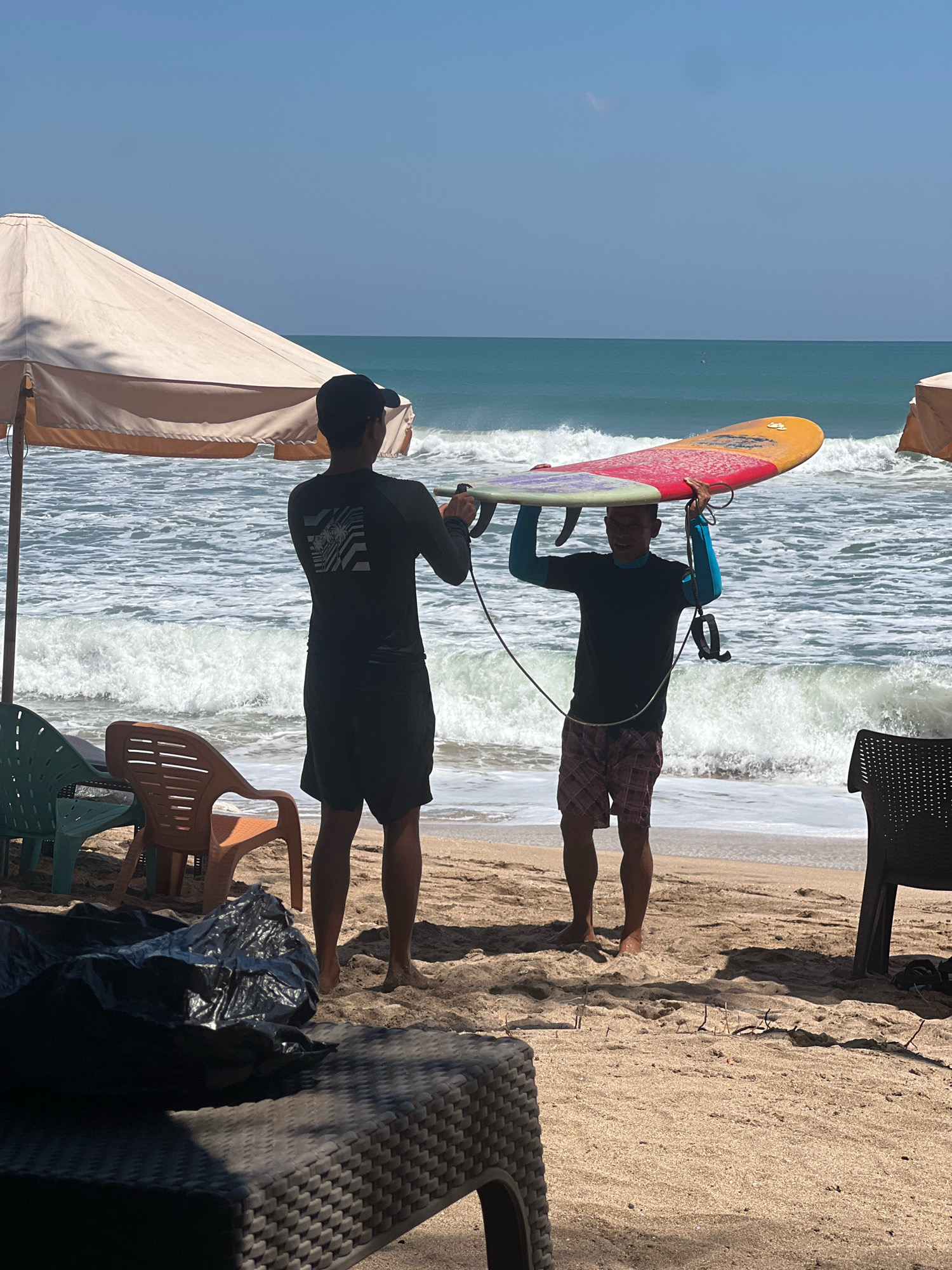 Two men are on a sandy beach in Kuta, Indonesia, near the ocean with waves. One man, wearing a black t-shirt and shorts, stands facing away from the camera, holding a surfboard above his head with the assistance of the second man. The second man, wearing a black t-shirt and patterned shorts, is facing the camera and also holding the surfboard. The surfboard is colorful, with red, yellow, and blue hues. In the background, the ocean is a deep blue with white waves. Umbrellas and chairs are visible on the beach, indicating a recreational area.
