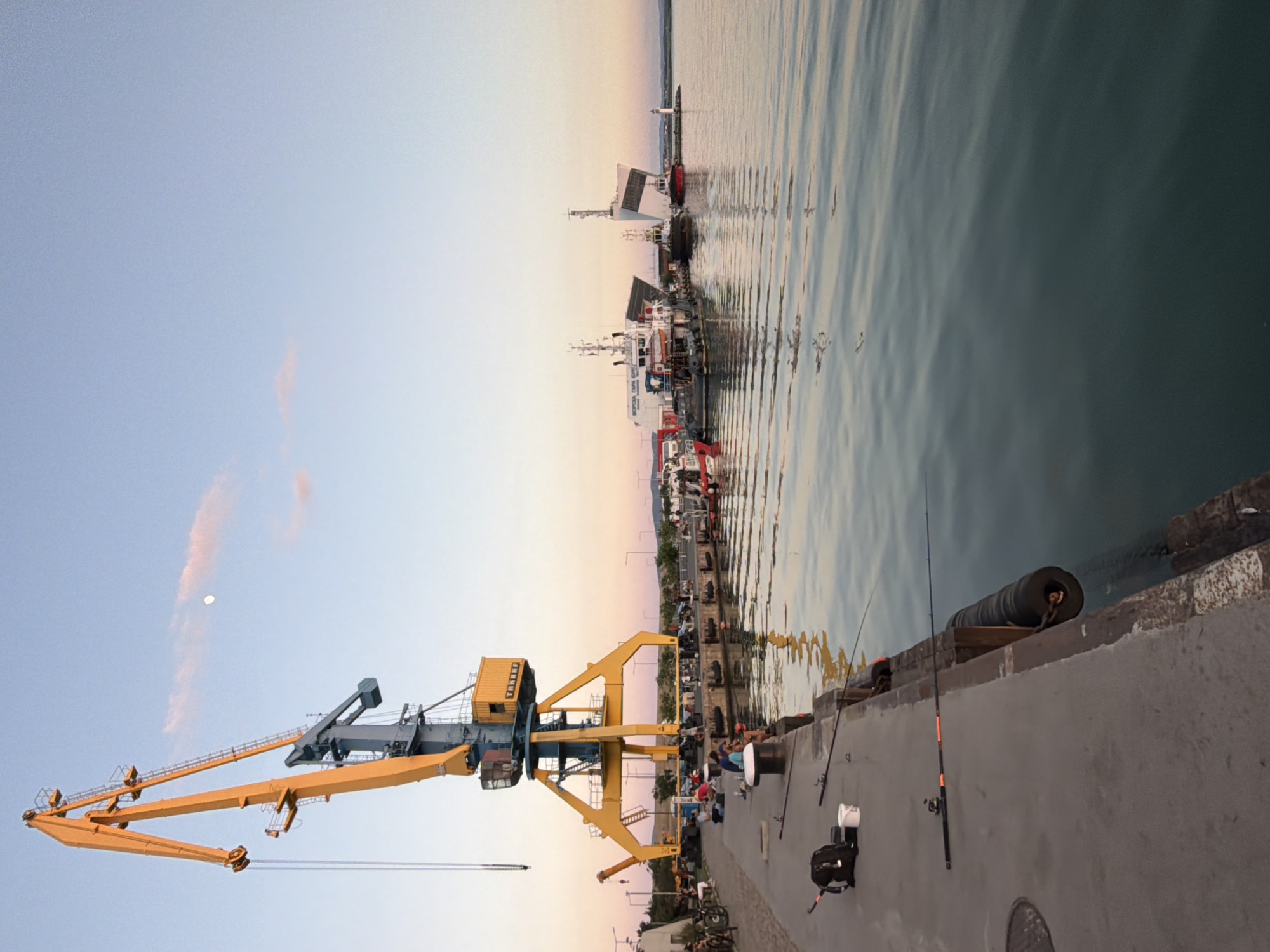 The image depicts a waterfront scene in Burgas, Bulgaria. A large yellow crane dominates the left side of the frame, standing on a concrete pier. Several people are fishing along the pier, their fishing rods extending towards the water. In the background, ships are docked along the harbor. The water is calm and reflects the colors of the sky, which ranges from blue to orange as it approaches the horizon.