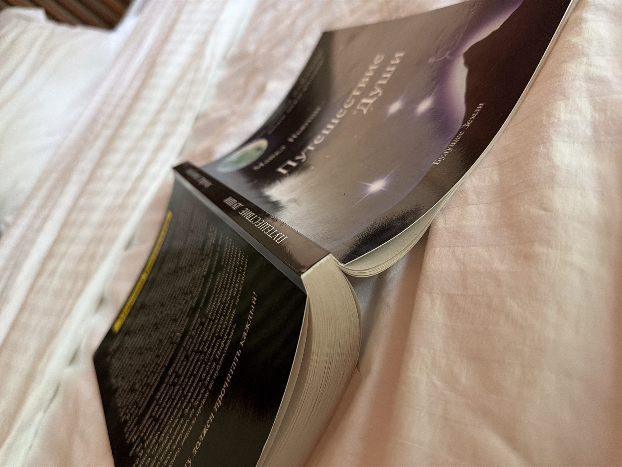 An open book lies on a pink textured surface, possibly a bedspread. The book's cover is dark with a starry, cosmic design and text in a light color. The title appears to be "Путешествие Души" (Journey of the Soul) and below it, "Будущее Земли" (Future of Earth). Part of the back cover is visible on the left, featuring a block of text and a yellow line at the top edge. The lighting suggests an indoor environment, and the surface appears soft and slightly wrinkled. There are no people or animals visible in the image, and the scene is static. The location is not identifiable as Ko Samui District, Thailand, or any specific city.