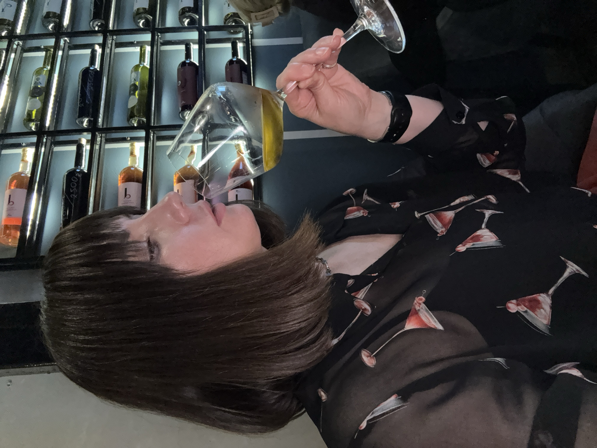 A woman with dark brown hair and bangs is holding a glass of amber liquid to her lips. She is wearing a black sheer blouse with a martini glass pattern and a silver necklace. The background shows a wine rack filled with bottles, suggesting a restaurant or bar setting. The scene appears to be indoors, with artificial lighting. The location is not identifiable as Izhevsk, Russia, from the image provided.