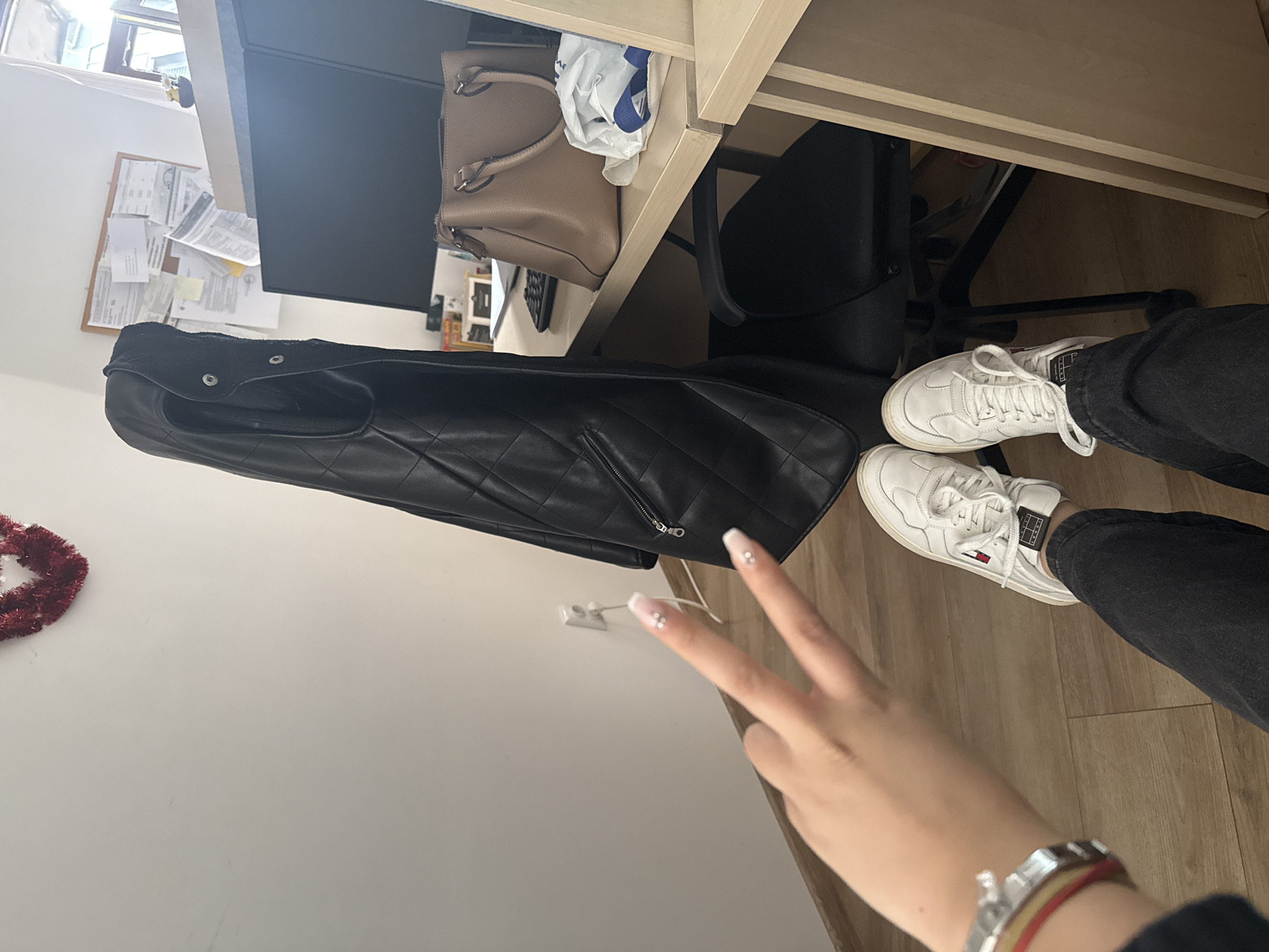 The image shows a first-person view of a person sitting at a desk. The person is wearing white sneakers, black jeans, and a silver wristwatch with a red band. They are holding up two fingers in a peace sign. On the desk is a black quilted handbag, a beige handbag, and a computer monitor. There is a black office chair and a corkboard with papers on the wall. The background is a plain white wall with a red garland in the corner. It is not possible to determine the exact location within Sofia, Bulgaria, from this image.