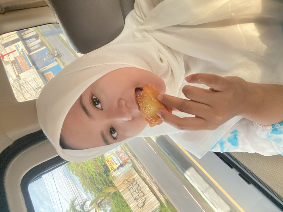 A woman in a white hijab sits in a car and eats a fried pastry. Her head is tilted to the side, and she looks at the camera with dark eyes. Her right hand holds the pastry to her mouth. Outside the car window, a street with buildings and foliage is visible. The car's interior includes a headrest and a light-colored dashboard.