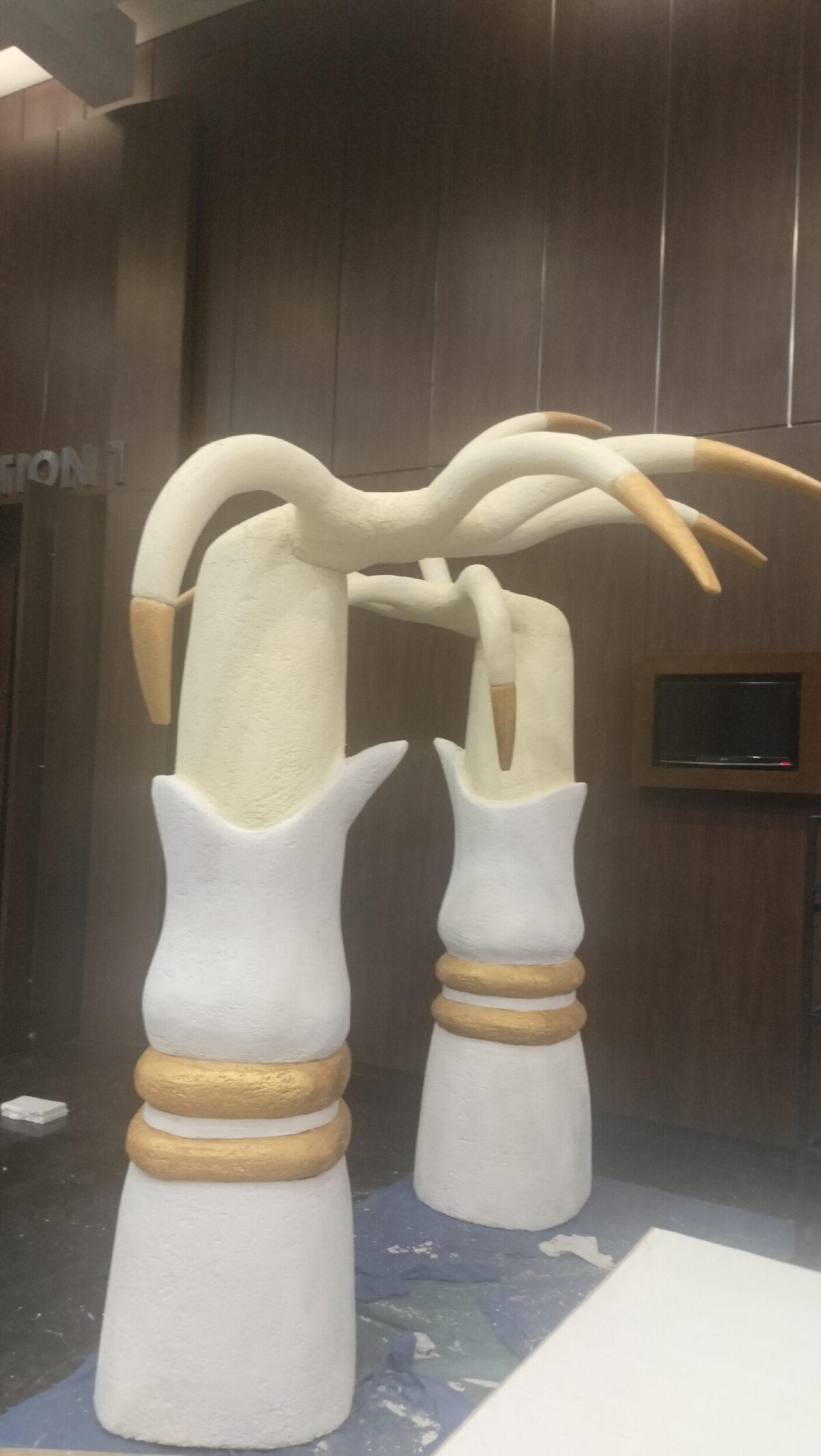 The image shows an indoor scene with two large, abstract sculptures in the foreground. The sculptures are primarily white and cream-colored with gold accents. They appear to be made of a textured material, possibly plaster or a similar composite. The left sculpture is taller than the right one, and both are wider at the base, tapering towards the top. Each sculpture has a flared opening at the base and two distinct gold rings around the lower portion of the cylindrical body. Above the main bodies, the sculptures branch out into curved, smooth forms that resemble stylized arms or branches. These upper sections are cream-colored and terminate in pointed, gold-tipped ends.
The sculptures are positioned on a dark blue cloth or tarp, with some white powder or debris scattered on the floor around them, suggesting they are either under construction, being assembled, or have recently been worked on.
In the background, a wall with vertical wooden paneling is visible. To the left of the sculptures, partially obscured by the left sculpture, is text that appears to read "ION 1". Further to the right in the background, a small, dark television screen is mounted within a wooden frame on the wall. The lighting in the scene is artificial and appears to be coming from overhead, casting soft shadows.
The overall setting suggests an indoor space, possibly a studio, workshop, or a public area where these sculptures are being prepared or displayed. There are no people visible in the image. The context of "City of Medan, Indonesia" is provided but not directly evident from the visual elements within the photograph itself.