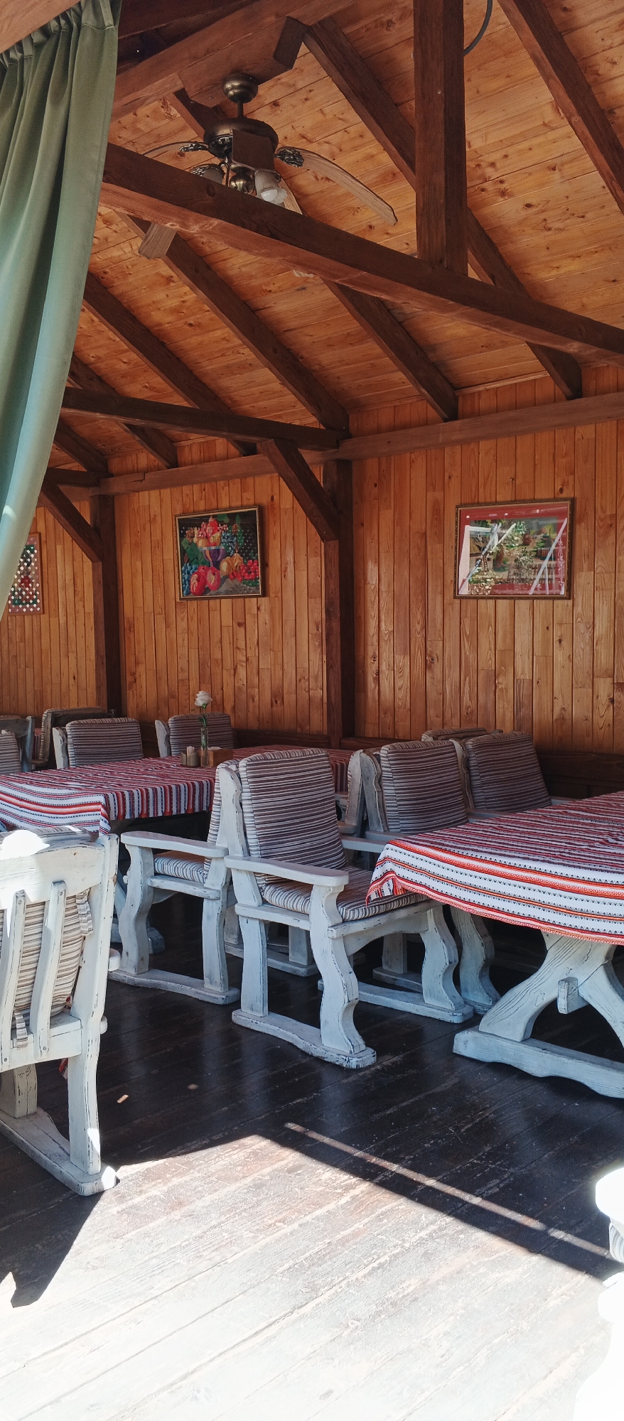 The image depicts an outdoor seating area, likely a restaurant or cafe, with a wooden roof and walls. A ceiling fan is visible, suggesting it is a warm climate. Tables are covered with striped cloths, and white wooden chairs with striped cushions are arranged around them. Two framed artworks are hanging on the wall, one of which appears to be a still life of fruits. The floor is made of dark wooden planks. Sunlight casts shadows across the floor, indicating it is daytime. The scene is tranquil and appears to be unoccupied by people. The location is identified as Mukachevo, Ukraine, though there are no specific visual cues in the image to confirm this.