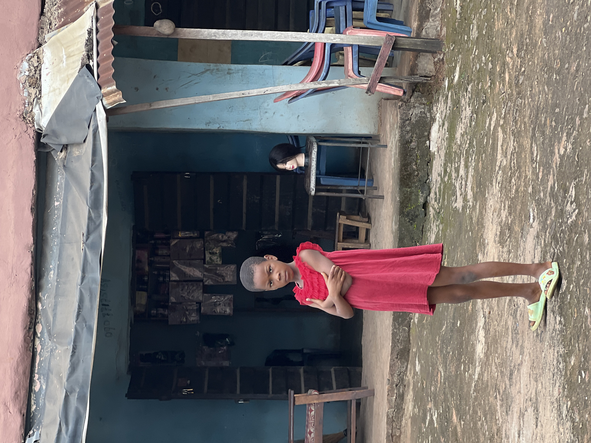A young girl stands with her arms crossed in front of a blue storefront in Nkpor, Nigeria. She is wearing a red dress and light green sandals. Behind her, products are displayed on shelves inside the store. To the right of the storefront, a mannequin head sits on a stool, and several colorful plastic chairs are stacked. The building appears to be made of concrete with a corrugated metal roof. The ground in front of the store is a mixture of concrete and dirt.