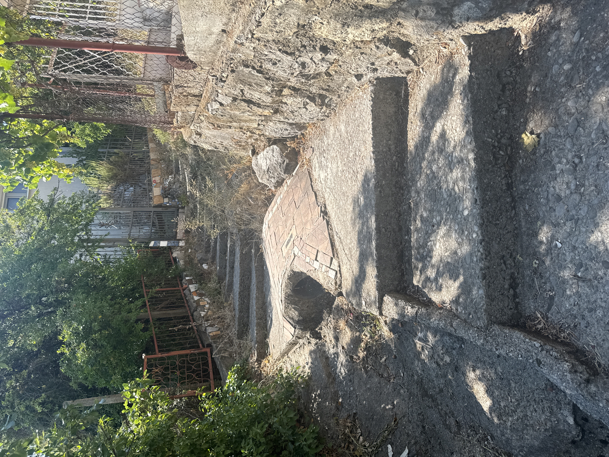 The image depicts an outdoor scene on a sunny day, likely in Brca, Montenegro. A set of uneven concrete stairs ascends from the foreground, covered in shadows from overhead foliage. The stairs are worn and appear to be part of an older structure, with visible cracks and debris. To the left of the stairs, dense green foliage and bushes are present. To the right, a rough stone wall borders the stairs, supporting a chain-link fence with red metal posts and a decorative red metal railing visible further up the path. Behind the fence, more greenery and what appears to be the side of a building can be seen. The upper portion of the image shows more trees and a rustic metal gate. The overall impression is one of an overgrown, somewhat neglected outdoor pathway.