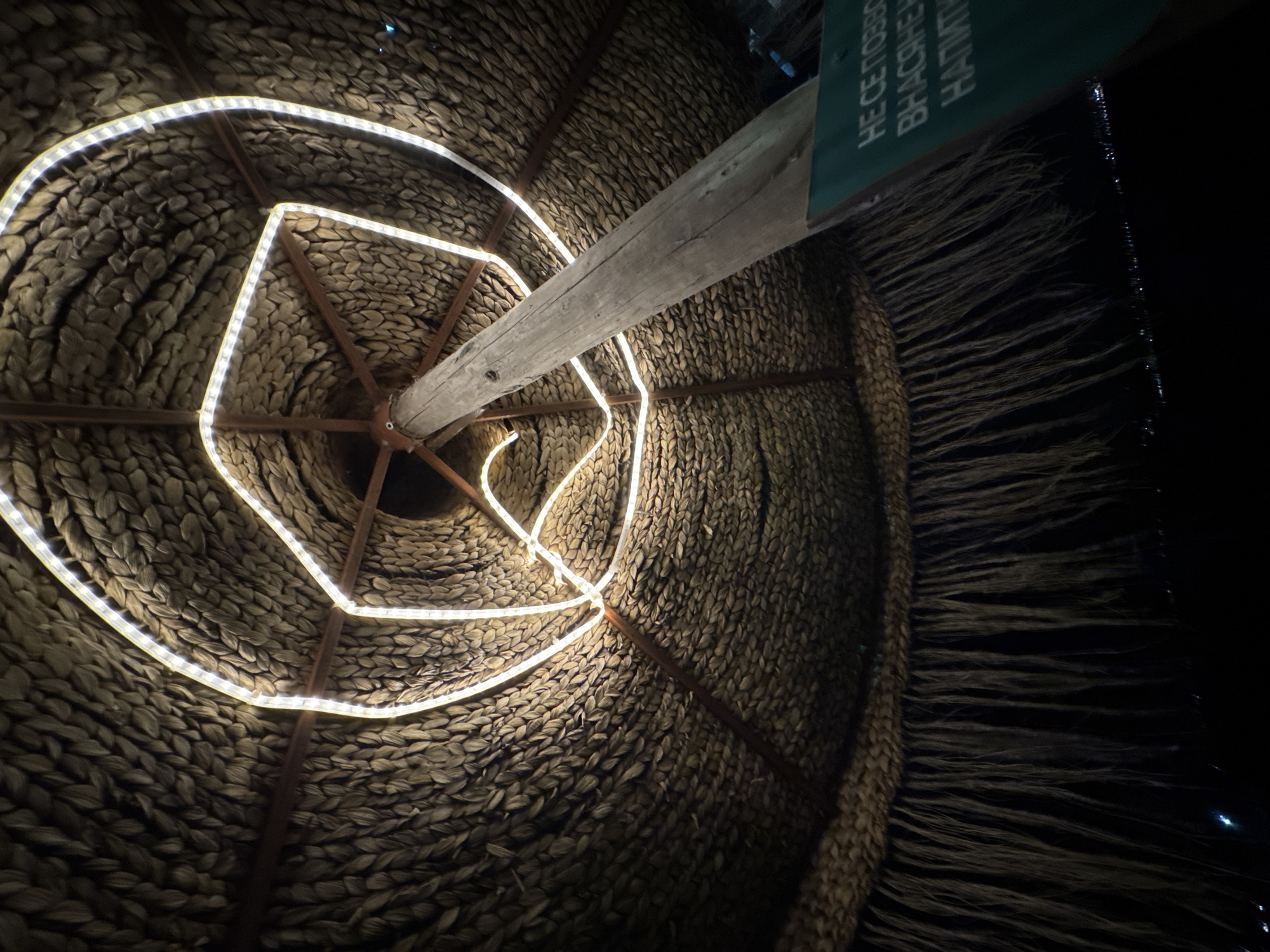 A tilted, low-angle shot depicts the interior of a thatched roof structure, possibly a palapa or similar construction, illuminated by LED strip lights. The roof's woven texture dominates the frame. A central wooden support pole extends diagonally across the image, with a sign attached to it on the right side. The background transitions to darkness on the right edge, suggesting an outdoor setting at night. This location appears to be within Varna, Bulgaria.
