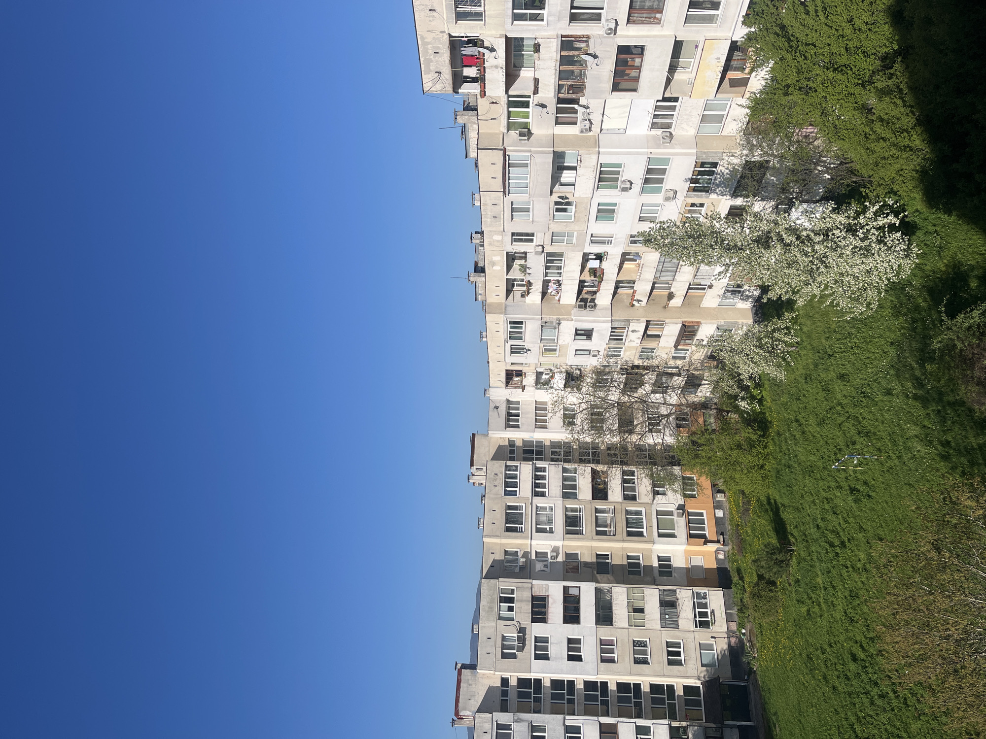 The image shows a high-angle view of a residential area in Sofia, Bulgaria. The scene features a clear blue sky on the left and two tall, Soviet-era apartment buildings on the right. Between the buildings is a grassy area with trees and flowering bushes. Some apartments have air conditioning units and clothes drying on balconies. The buildings appear to be well-maintained, although the paint is faded in places. The time of year appears to be spring.